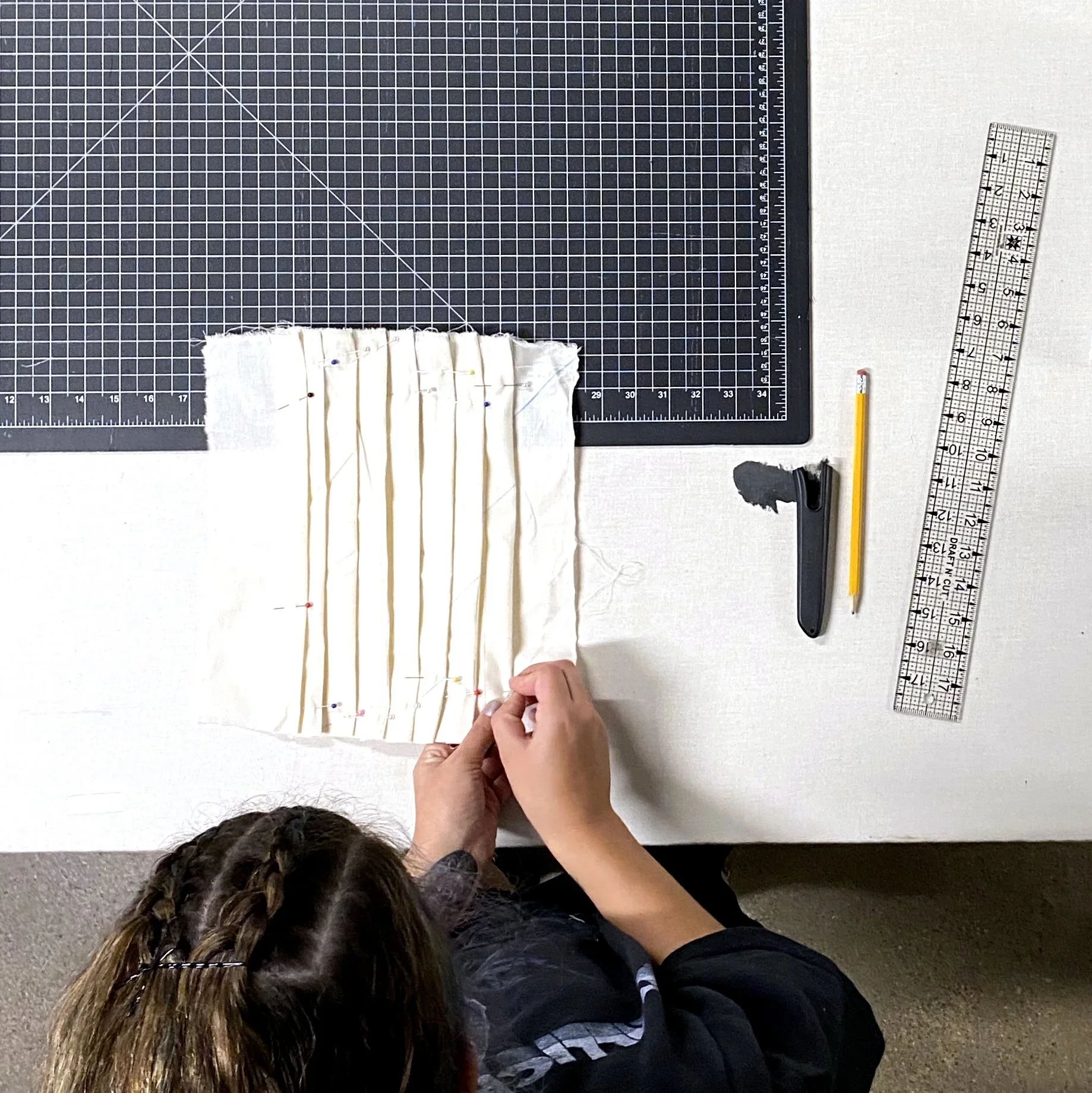 Person sewing fabric with pins on a table, surrounded by rulers and cutting mat.