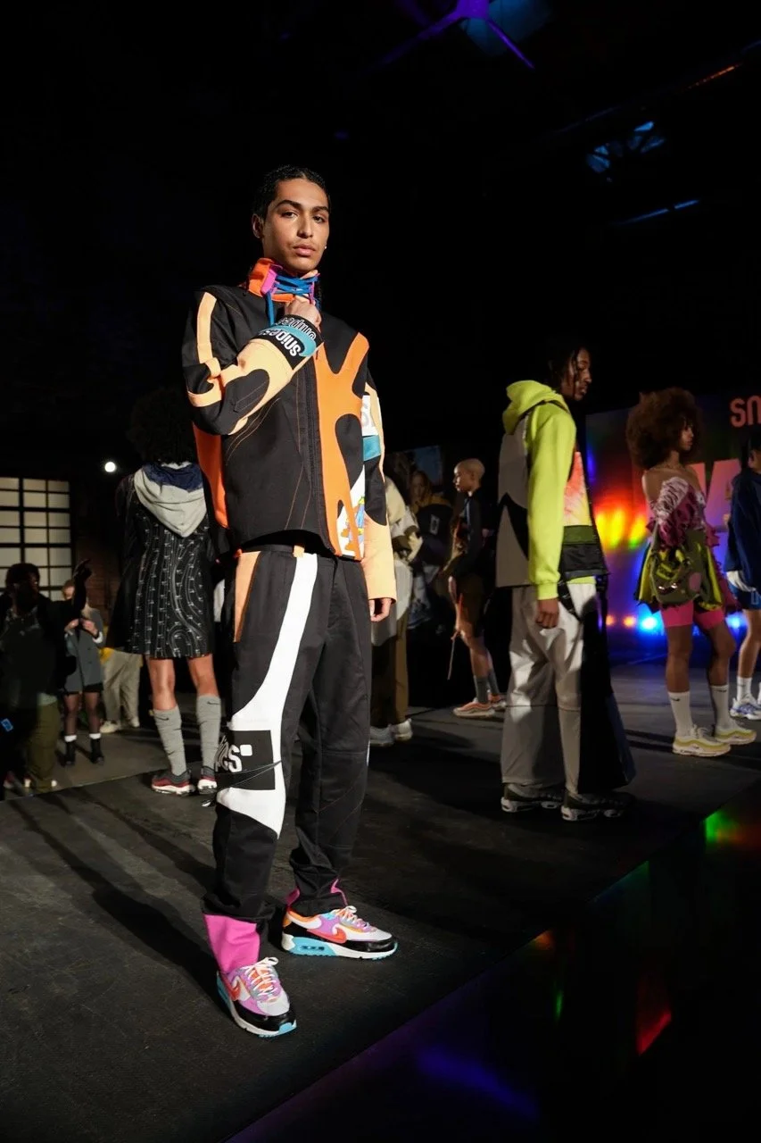Models on a runway at a fashion show, wearing colorful athletic and streetwear outfits, with a dark background and stage lighting.