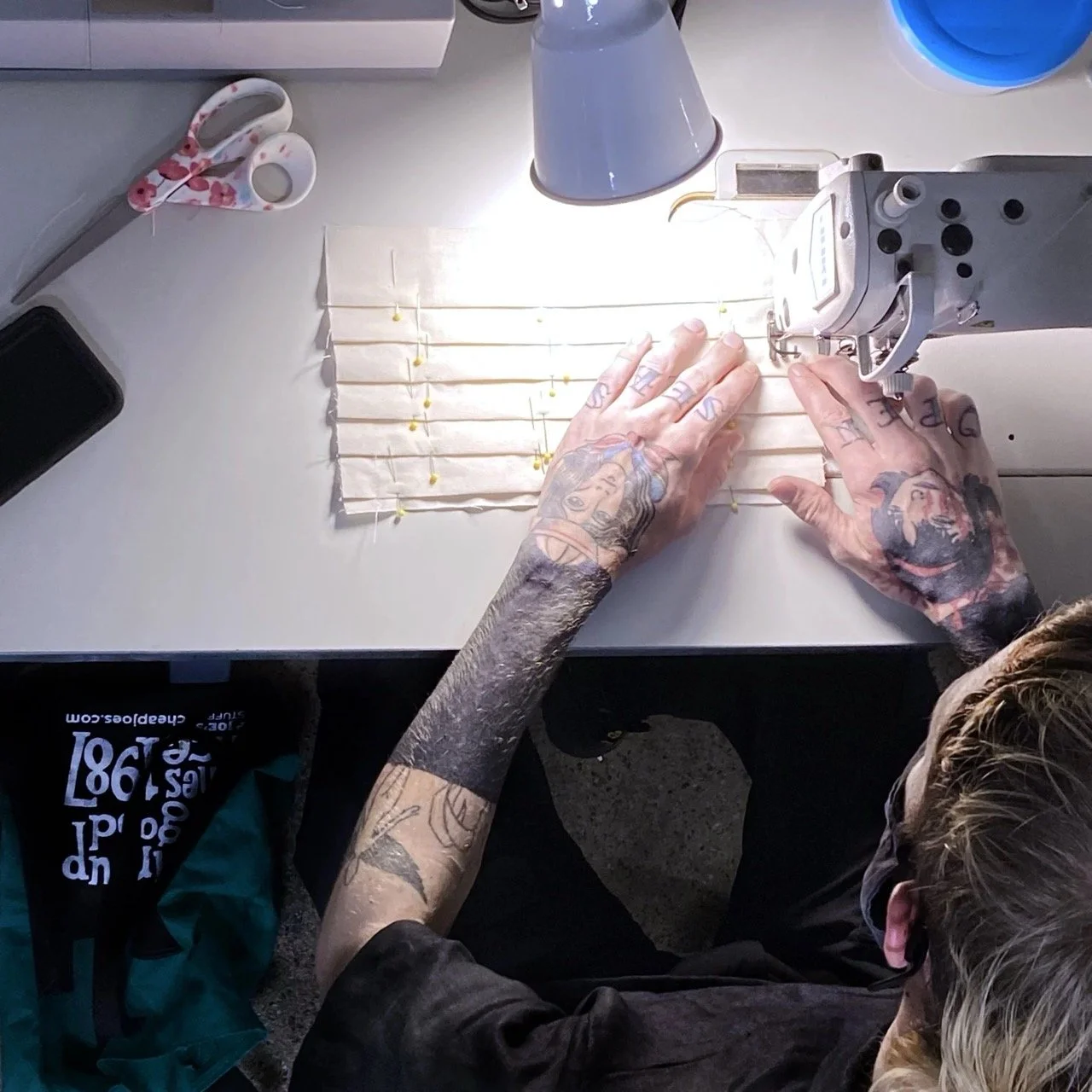 A person with tattooed arms working on a sewing machine, stitching a piece of fabric or paper, with various sewing tools and a lamp on the desk.