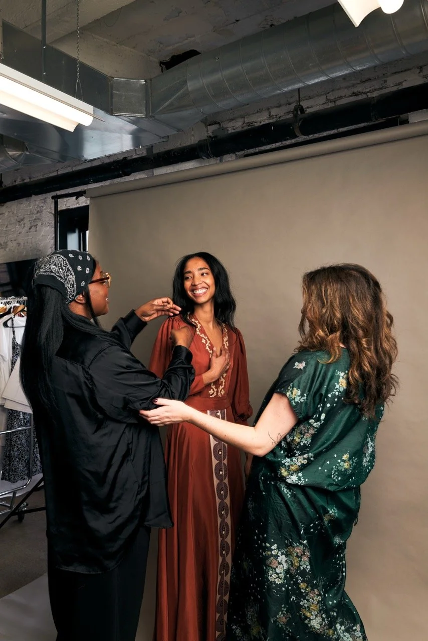A woman getting her photos taken, surrounded by two women assisting her in a studio with an industrial ceiling and a beige backdrop.