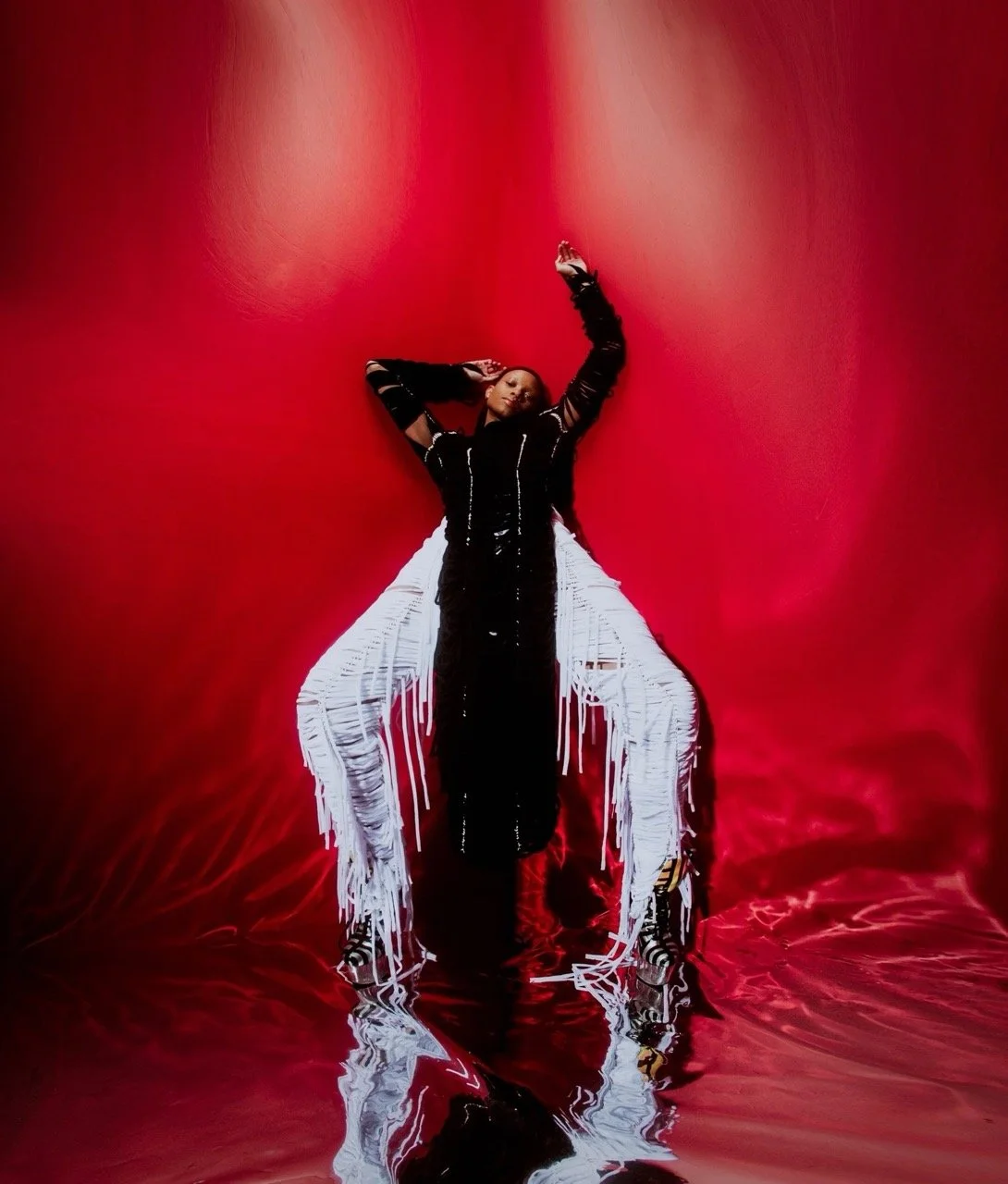 A person wearing a black outfit with white fringe stands against a red backdrop, striking a dramatic pose with one arm raised and the other hand behind their head.