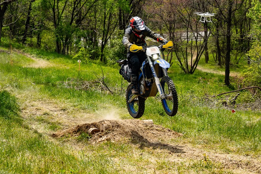 Motorcycle rider jumping off a dirt mound on a grassy trail in a wooded area, with a drone flying nearby.