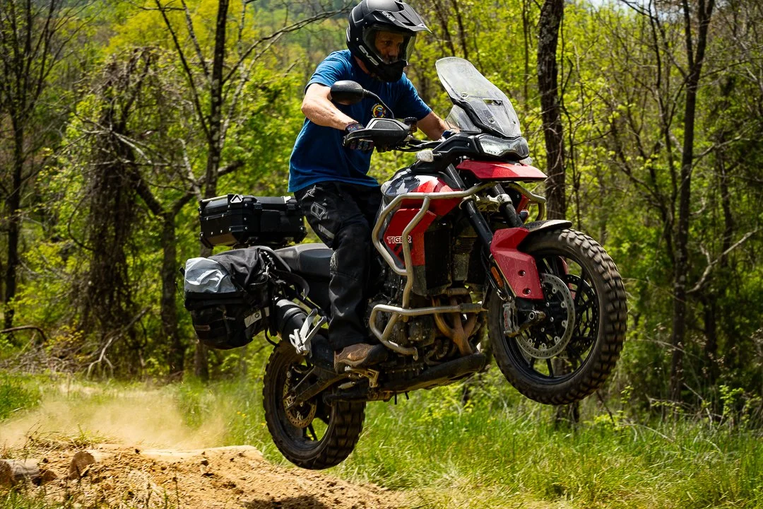 Motorcycle rider in a blue shirt and black helmet jumping on a dirt trail surrounded by trees.