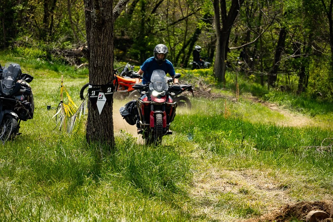 Two motorcyclists riding through a wooded trail with grass and trees, with parked motorcycles and a bicycle nearby.