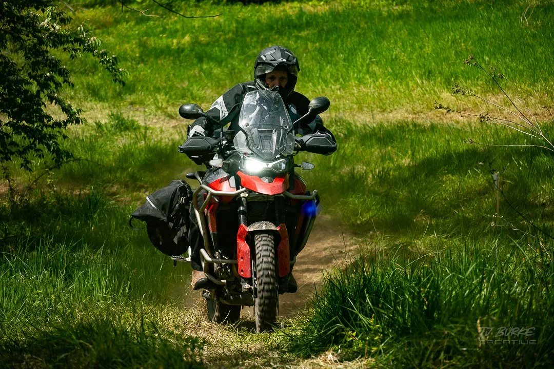 Motorcycle rider wearing a helmet and riding gear on an off-road trail surrounded by green grass and trees.