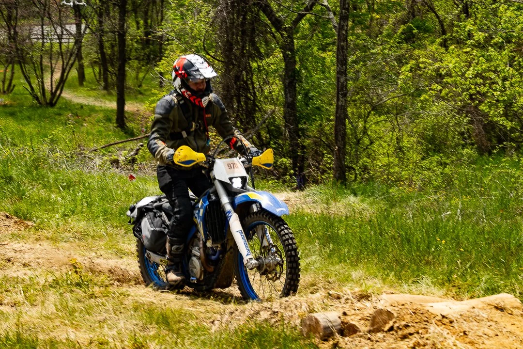Motorcyclist riding a dirt bike on a wooded trail with green grass and trees.