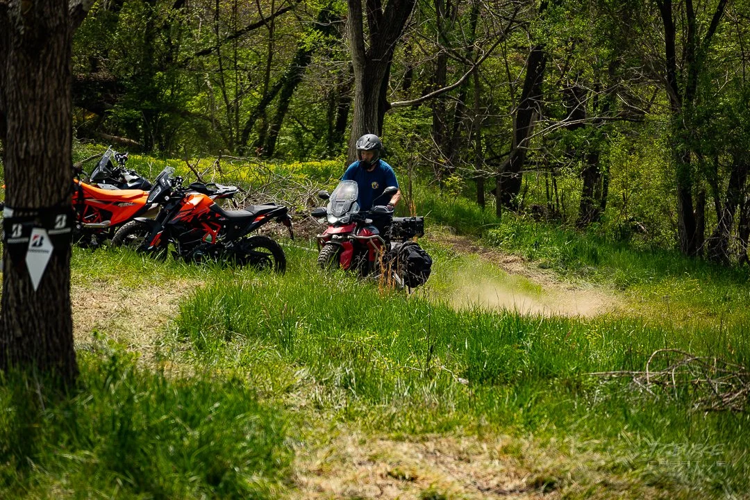 A person standing next to three parked adventure motorcycles on a forest trail in a green wooded area.
