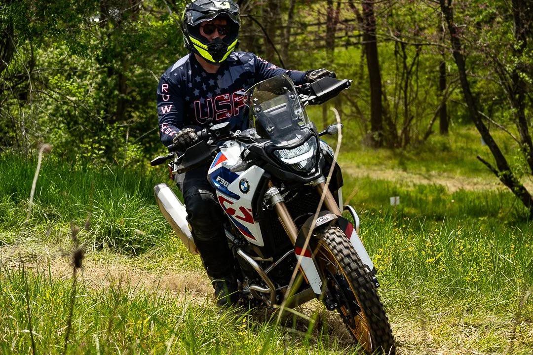 Motorcyclist riding an off-road BMW bike through a grassy trail surrounded by trees.