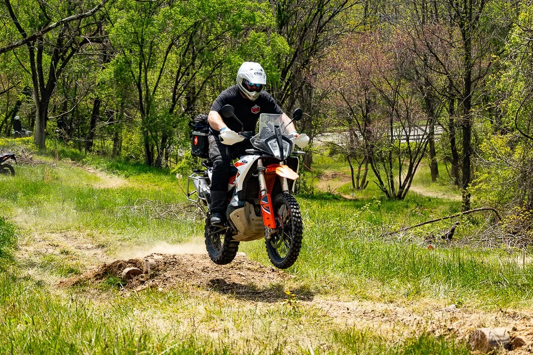 Motorcycle rider wearing a white helmet and black riding gear jumping over a dirt mound on a trail surrounded by green trees.