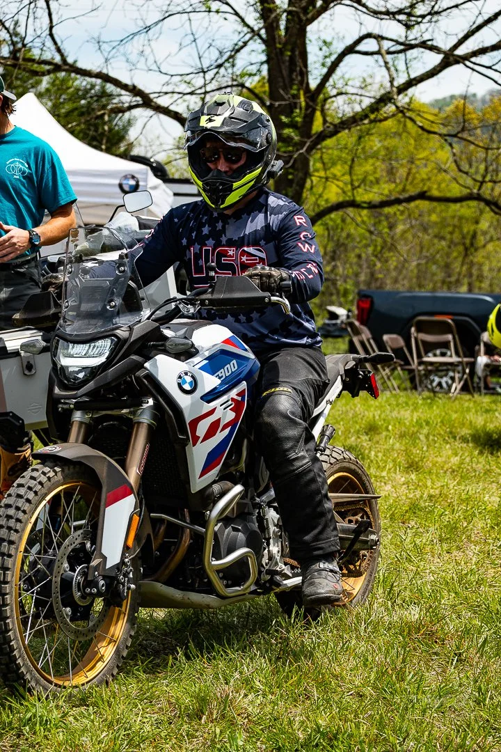 Person wearing a motorcycle helmet and riding gear sitting on a BMW motorcycle with a white, blue, and red racing design, outdoors on a grassy area with trees and tents in the background.