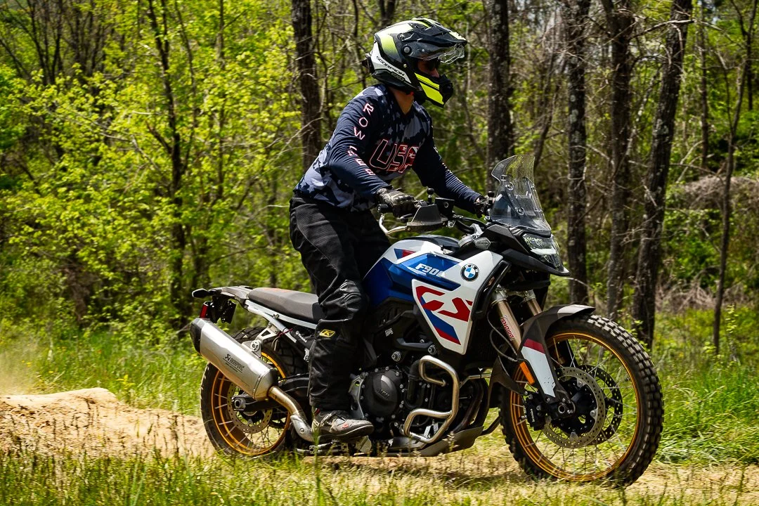 A person wearing a helmet and riding gear on a BMW off-road motorcycle in a forested area with green trees and grass.