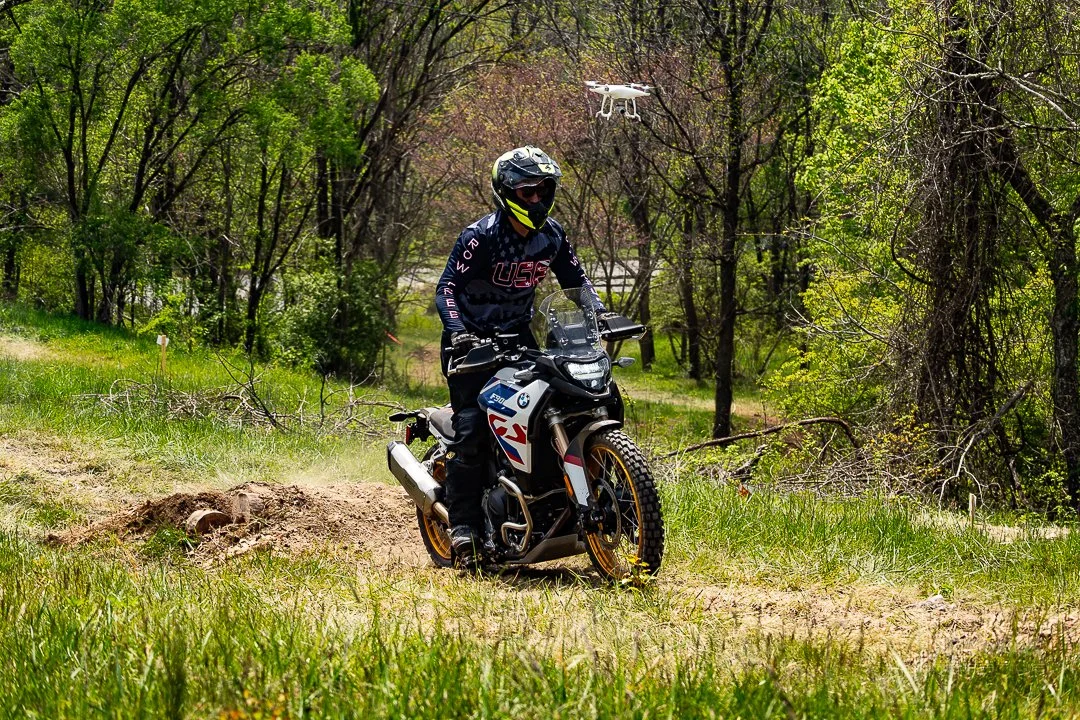 A person riding a BMW off-road motorcycle through a grassy trail in a wooded area, equipped with a helmet and riding gear, with a drone flying overhead.