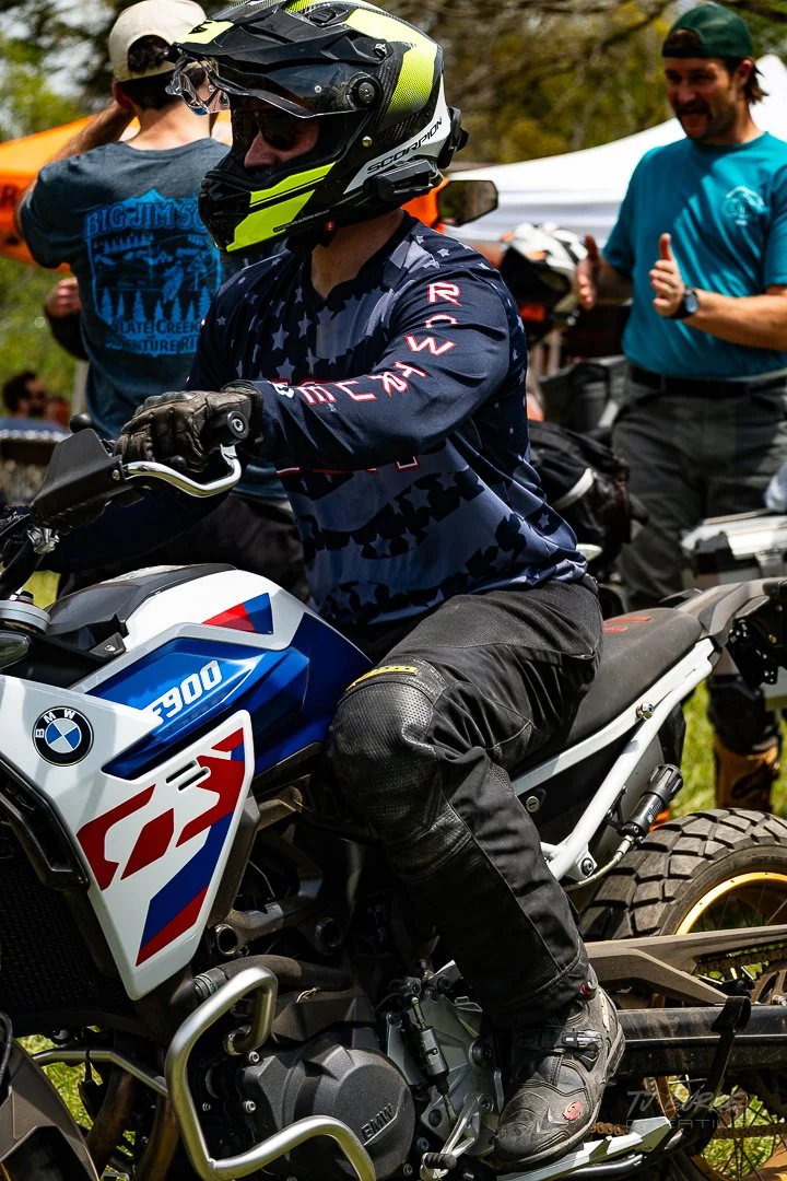 A person wearing a helmet and riding gear on a BMW GS motorcycle at an outdoor event, with people and tents in the background.