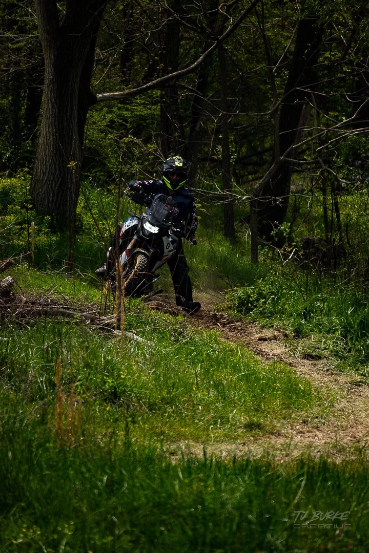 Person in black riding gear on a motorcycle through a forest trail surrounded by trees and green foliage.
