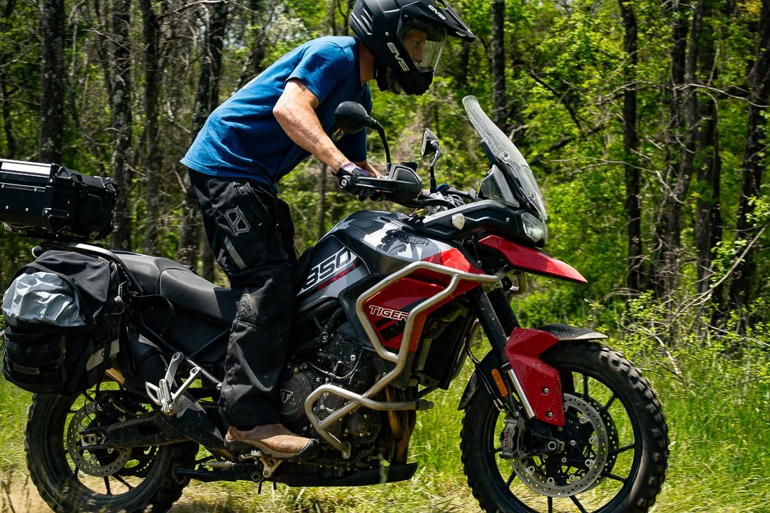 A person riding an adventure motorcycle on a dirt trail through a forest, wearing a helmet and riding gear.