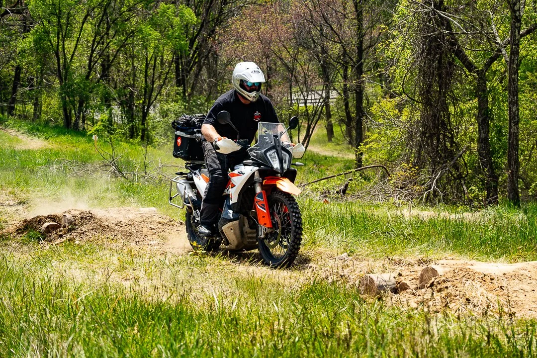 A person riding a dual-sport motorcycle on a dirt trail in a green wooded area.