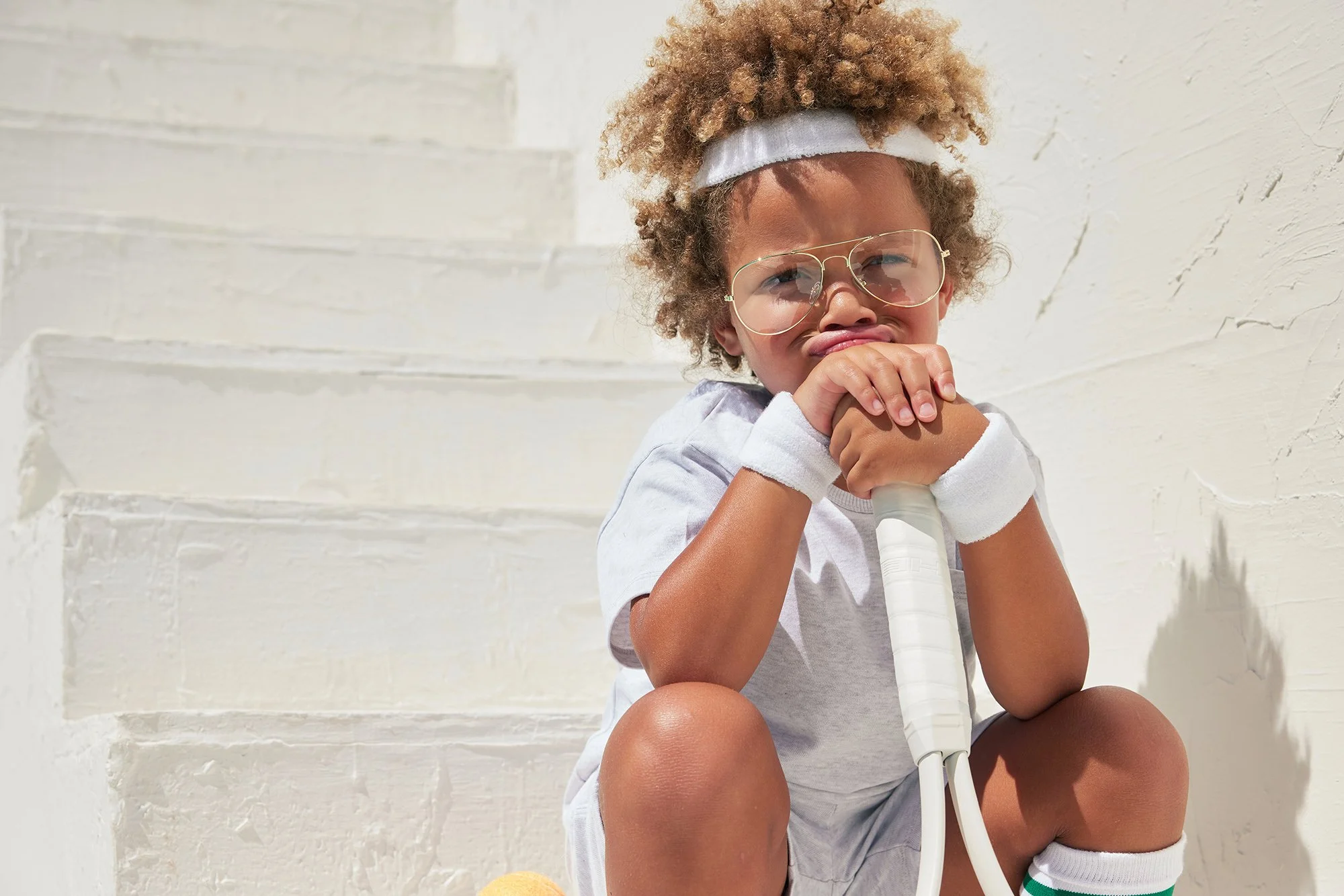 A young child with curly hair, wearing glasses and a white headband, sits against a white textured wall. The child is making a funny face by scrunching up their nose and has their hands clasped together near their mouth. They are wearing a light-colo