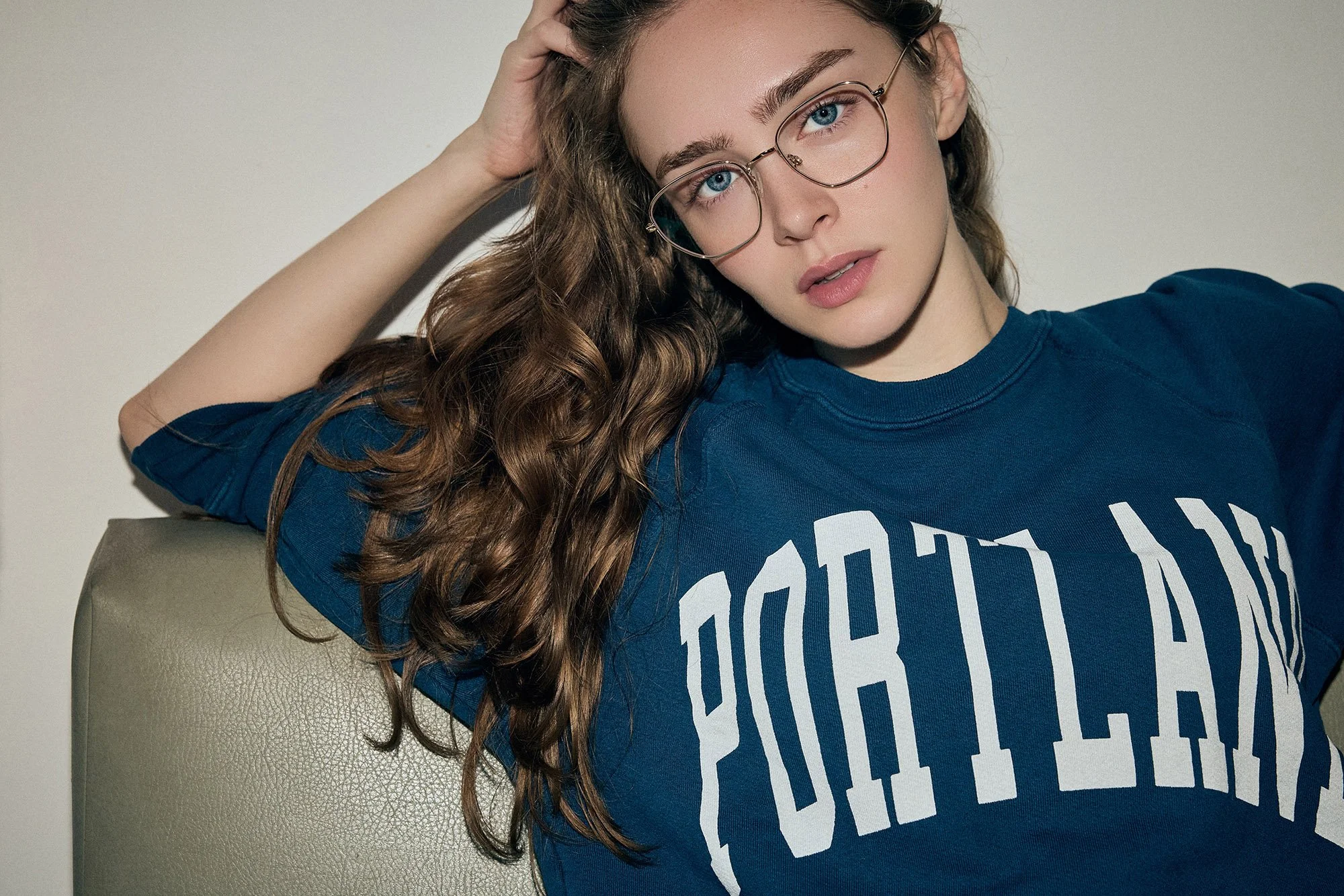 Fashion photography by Don Diaz — A young woman with long, wavy red hair and blue eyes, wearing glasses and a blue T-shirt that says 'PORTLAND' in white letters, sitting on a light-colored olive sofa against a plain white background.