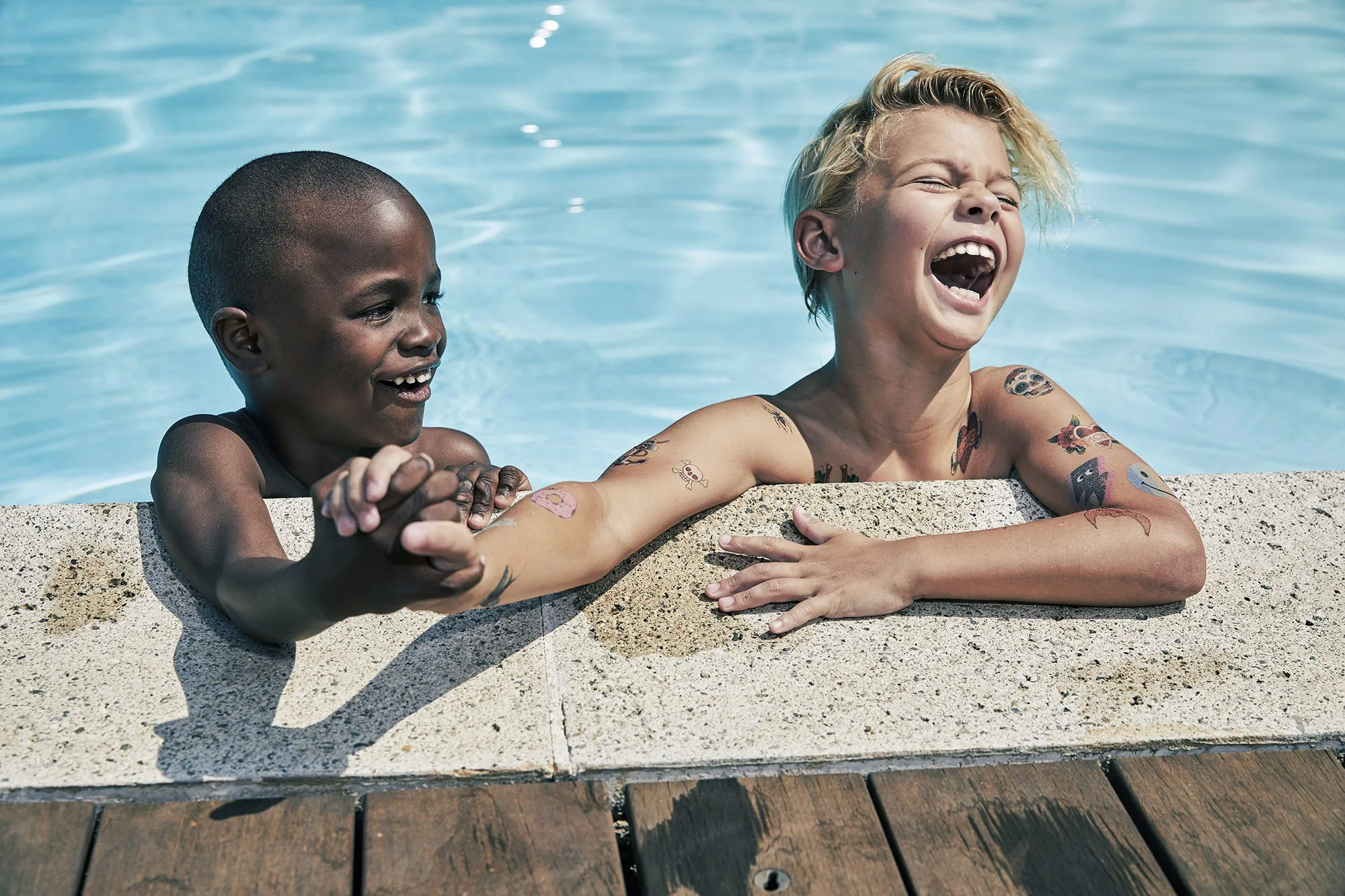 Two children smile and laugh as they hold hands over the edge of a swimming pool, with one resting on the pool's ledge and the other leaning in from the side. The pool water is clear and blue, and the children have tattoos and are enjoying a sunny da