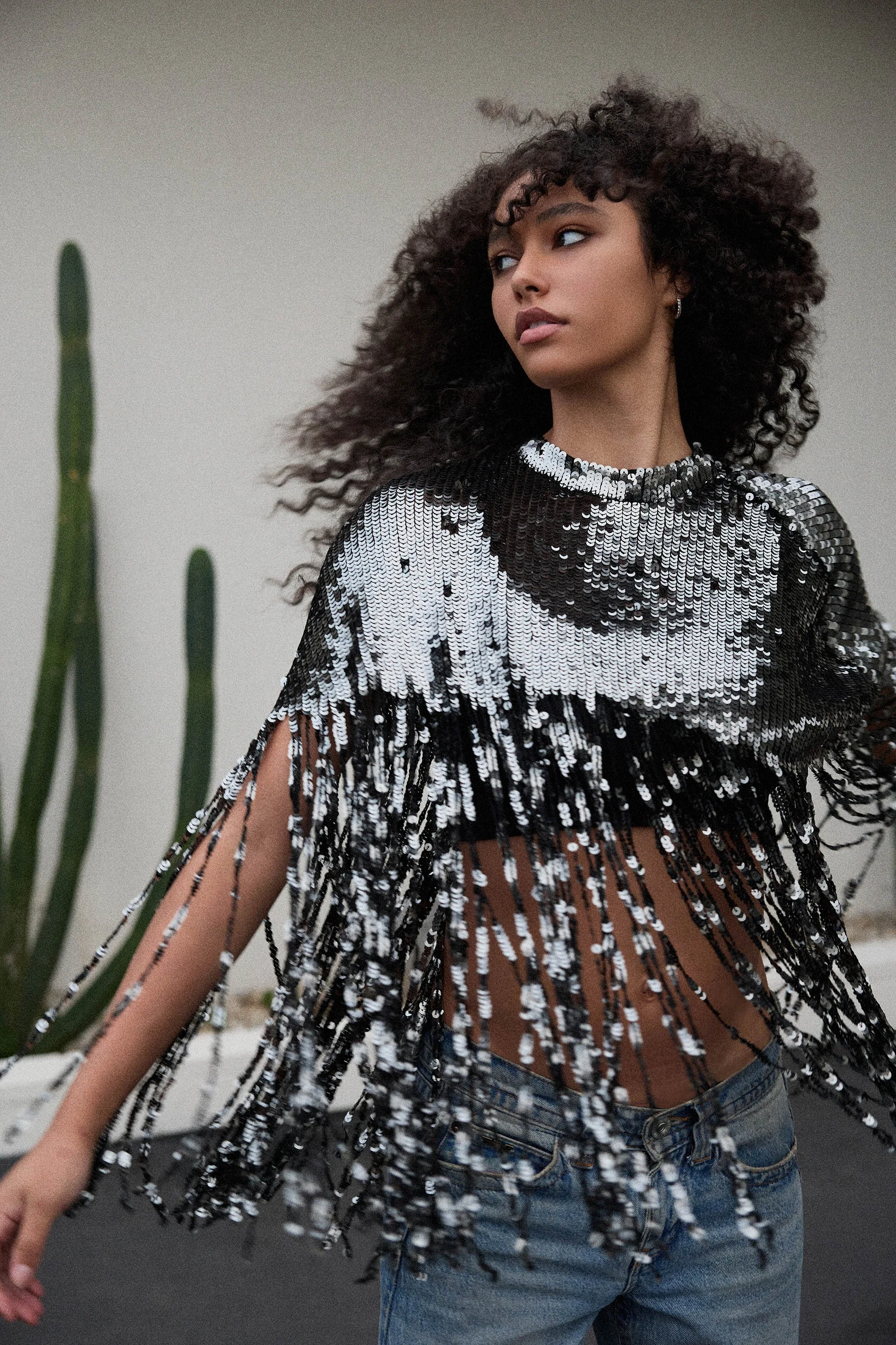 A young woman with curly hair wearing a black and white sequined top with fringe details, and jeans, standing near a cactus plant against a plain background.