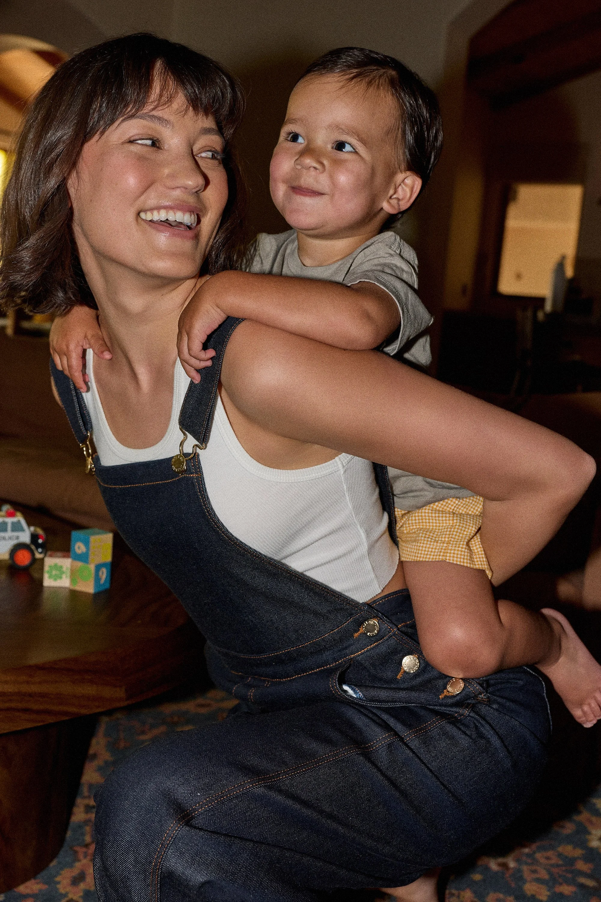 A woman with short brown hair and a white tank top is smiling while carrying a young boy on her shoulders. The boy has short dark hair, wears a gray t-shirt and yellow checkered shorts, and looks at the woman with a smile. The scene appears to be ind
