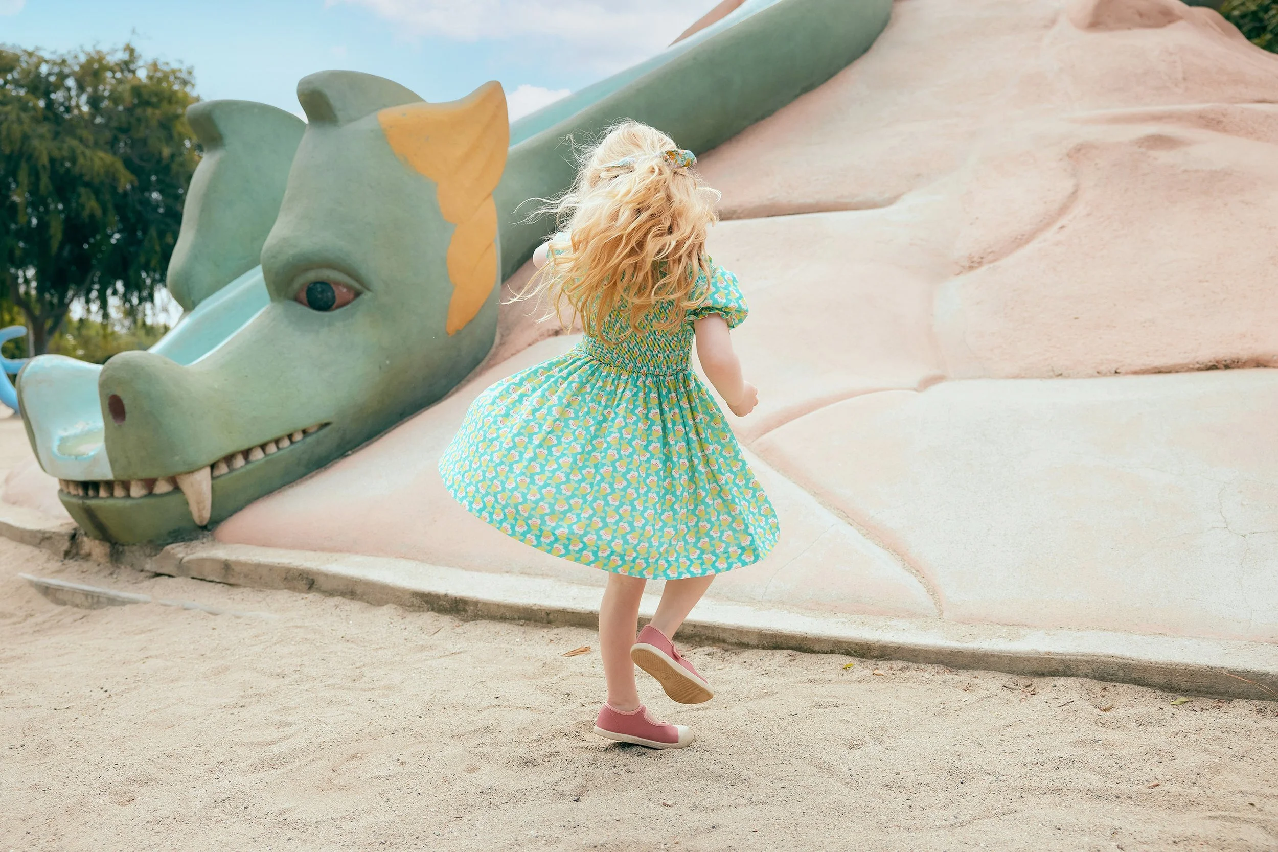 A young blonde girl in a patterned dress and pink shoes is playing near a large cartoon-like dragon sculpture outdoors, with trees and a blue sky in the background.