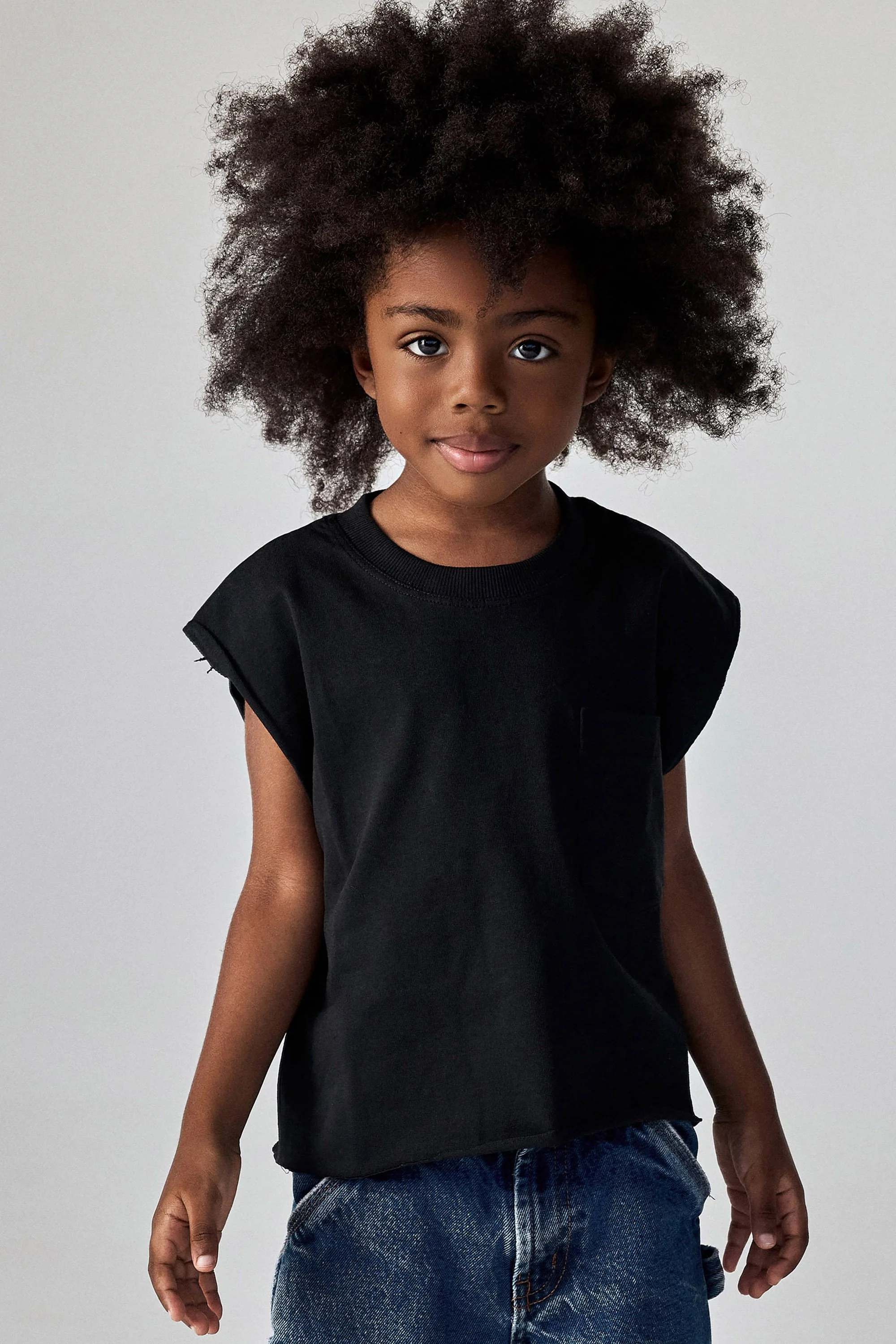 Young boy with big curly hair wearing a black T-shirt and jeans, standing against a light gray background.