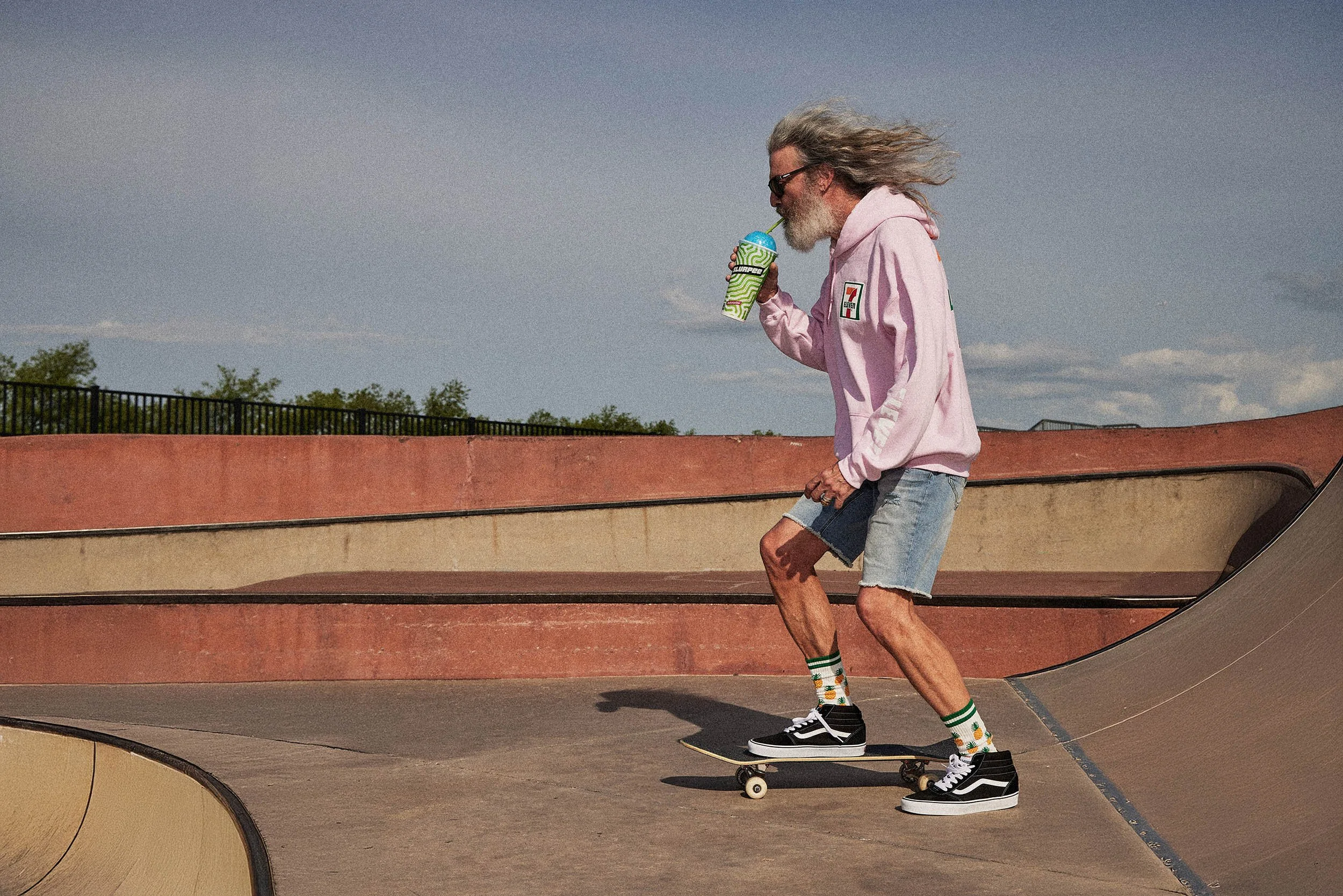 An older man with long gray hair and a beard skateboarding at a skate park. He is wearing a pink hoodie, denim shorts, black sneakers, and colorful socks. He is drinking from a slurpee cup with a straw while balancing on his skateboard.