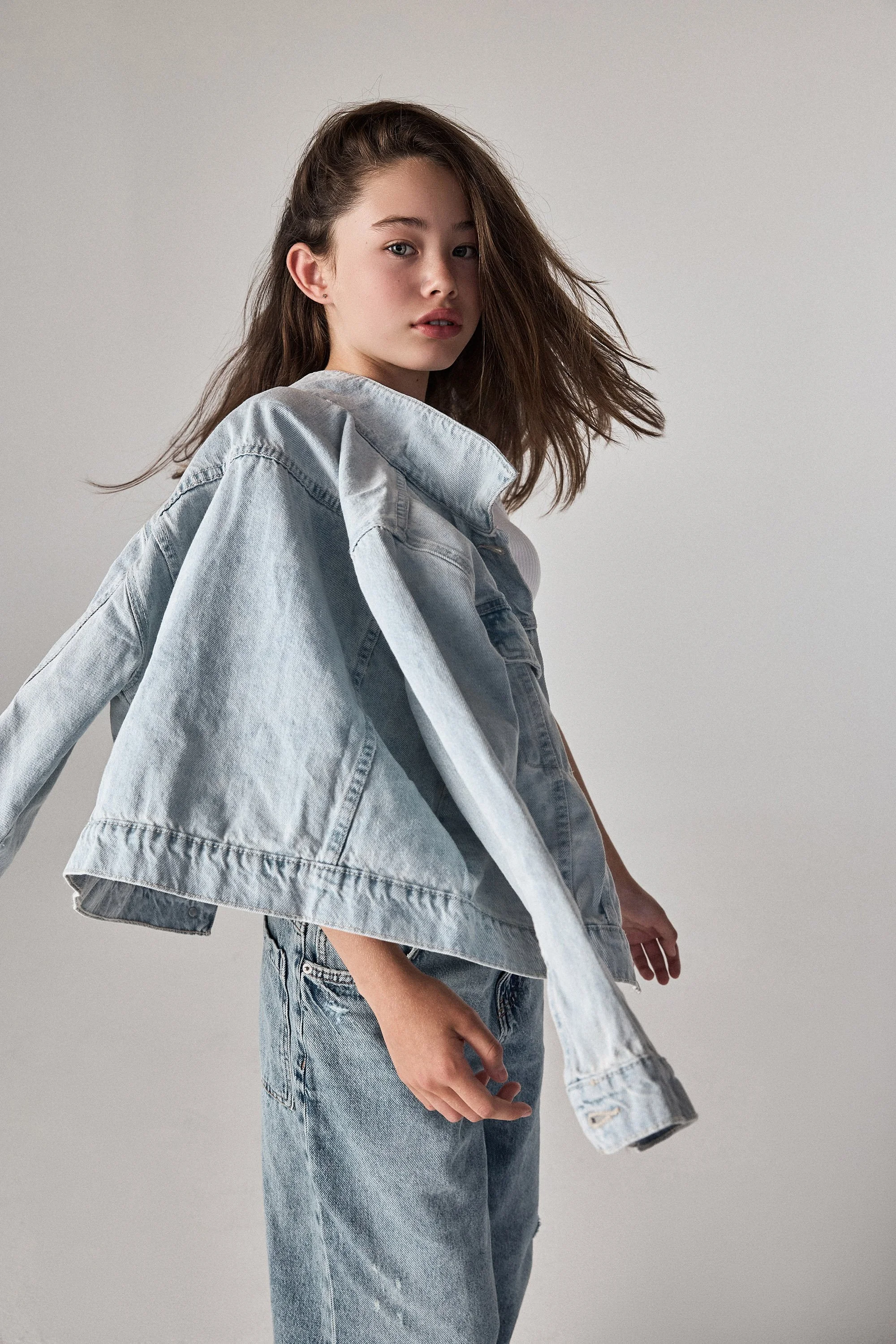 A tween girl  wearing a light denim jacket and jeans, standing against a plain light gray background. Her hair is blowing in the air from her movement.