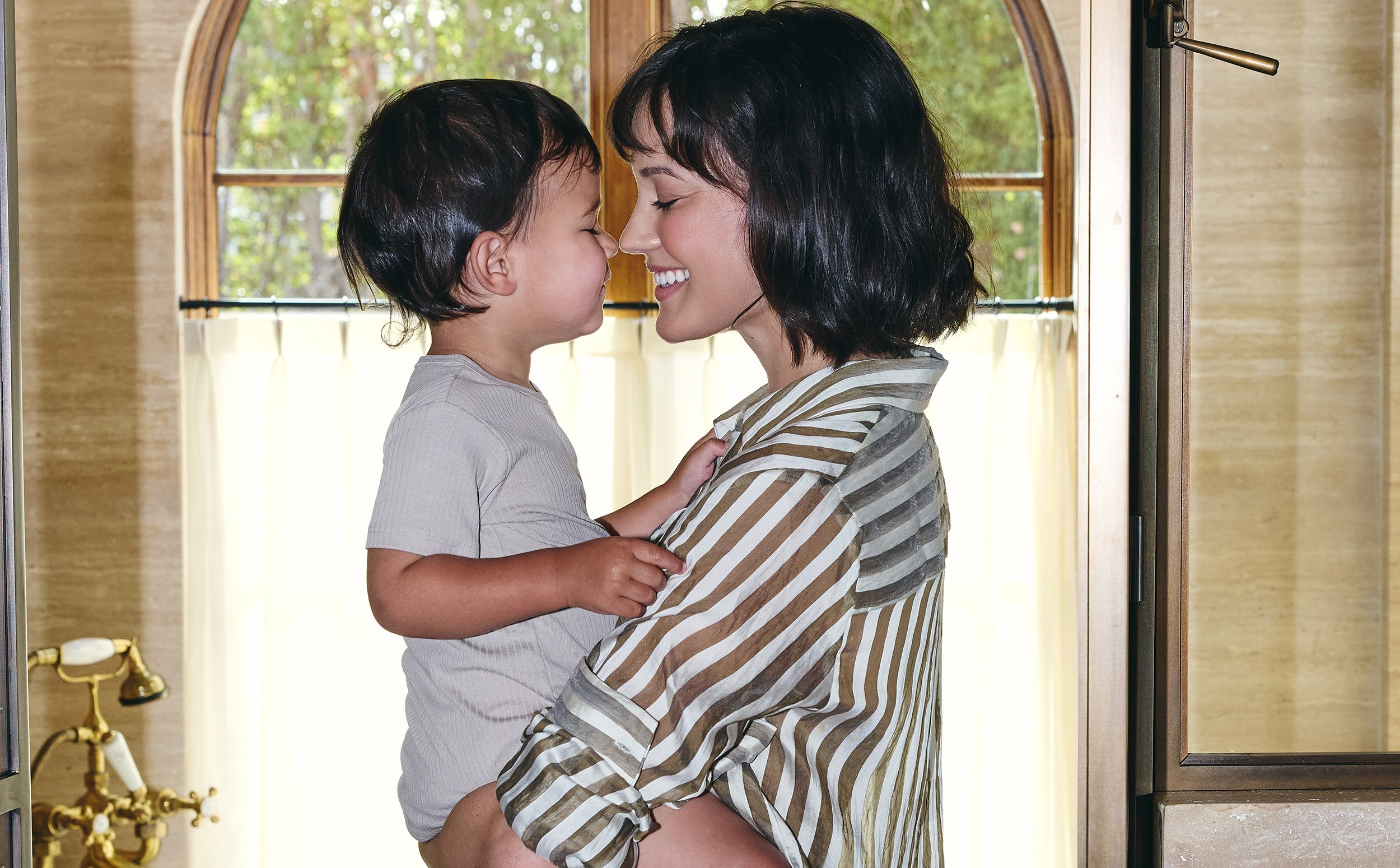 A woman and a young boy are standing close together inside a house near a door, touching their noses and smiling with eyes closed. The woman has short black hair and is wearing a striped shirt, while the boy has dark hair and is wearing a light gray 