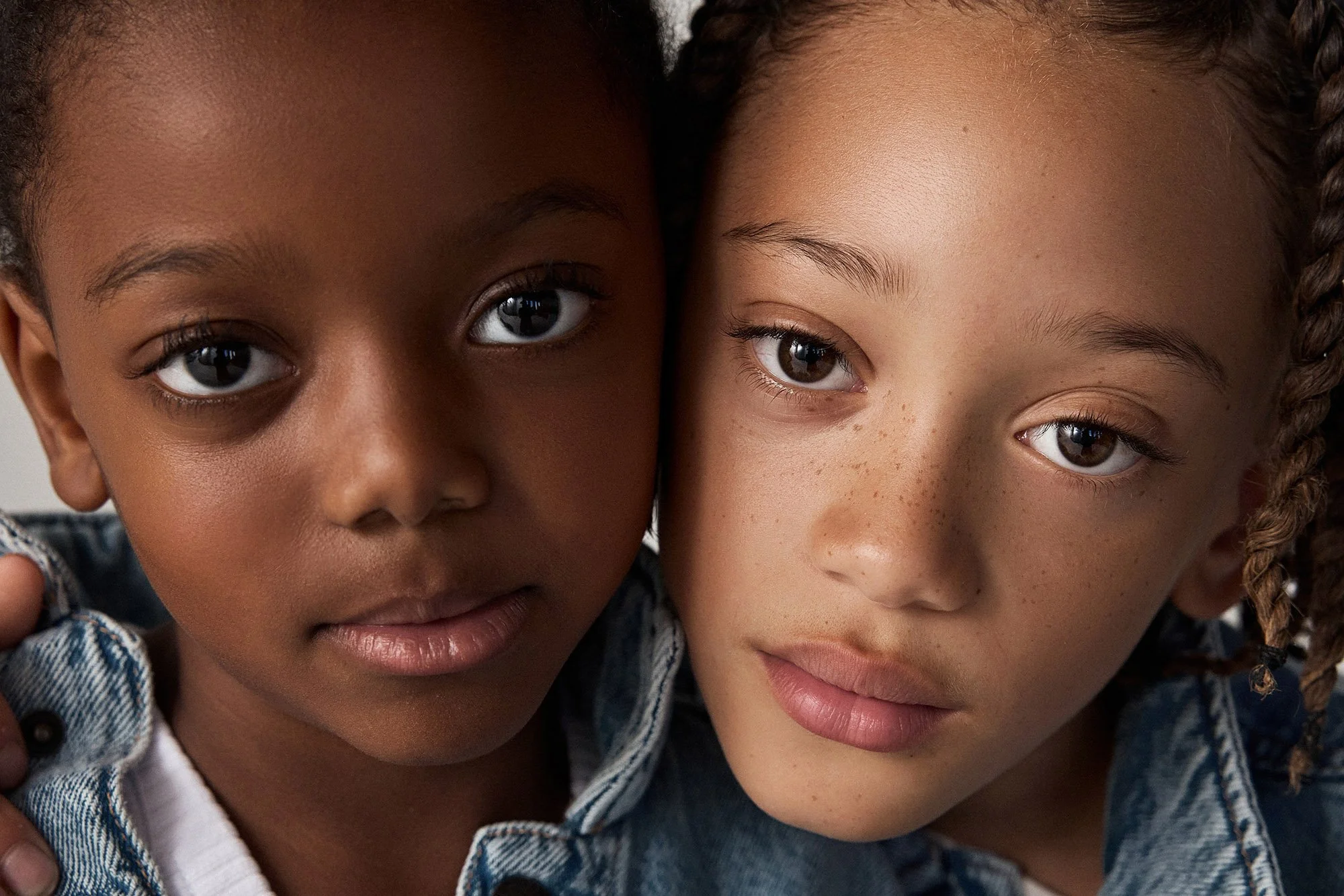 A close-up portrait of a girl and boy, one with dark skin and the other with light skin, both with brown eyes and braided hair, wearing denim jackets.