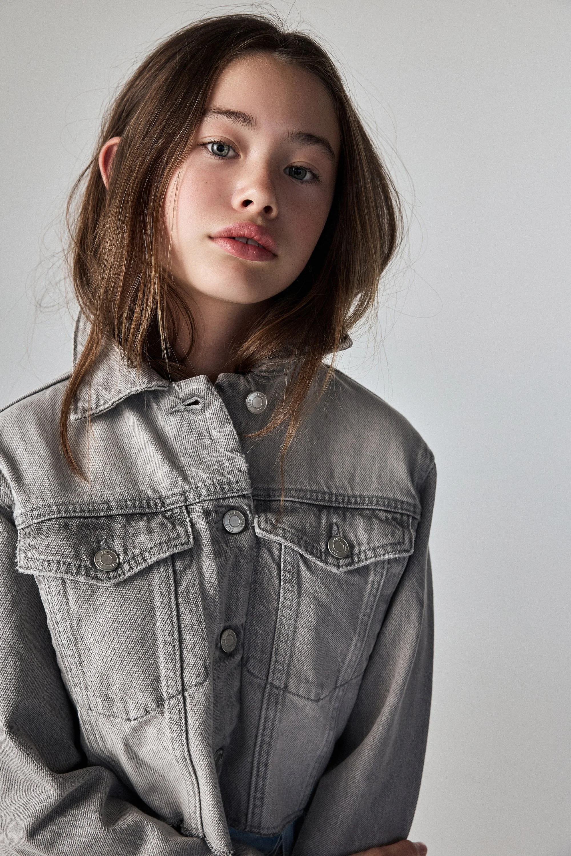 A young lady with brown hair and fair skin wearing a gray denim jacket against a plain, light-colored background.