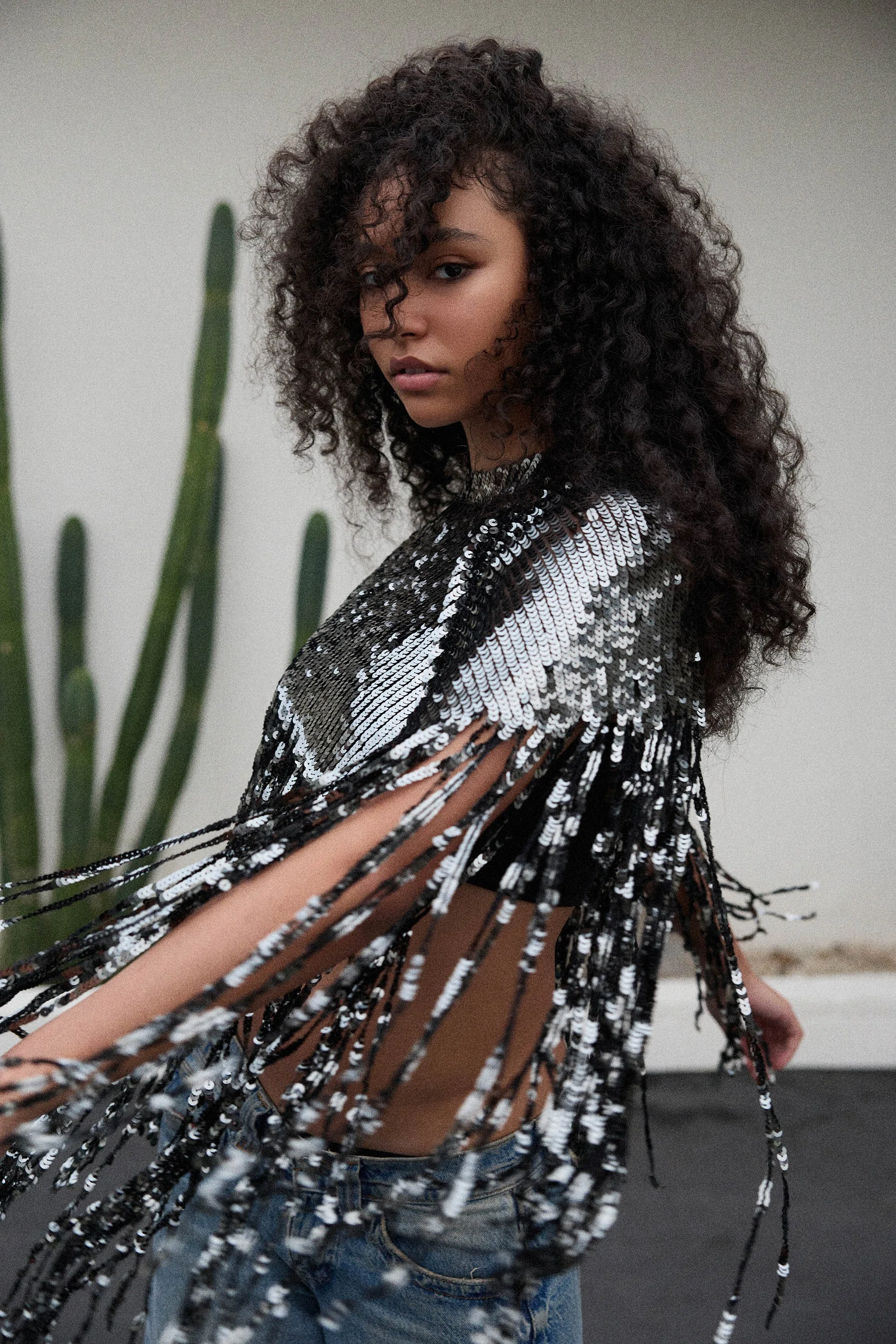 A young woman with curly hair wearing a black and silver sequined top and blue jeans stands against a neutral background with tall green plants. She looks at the camera with a confident expression.