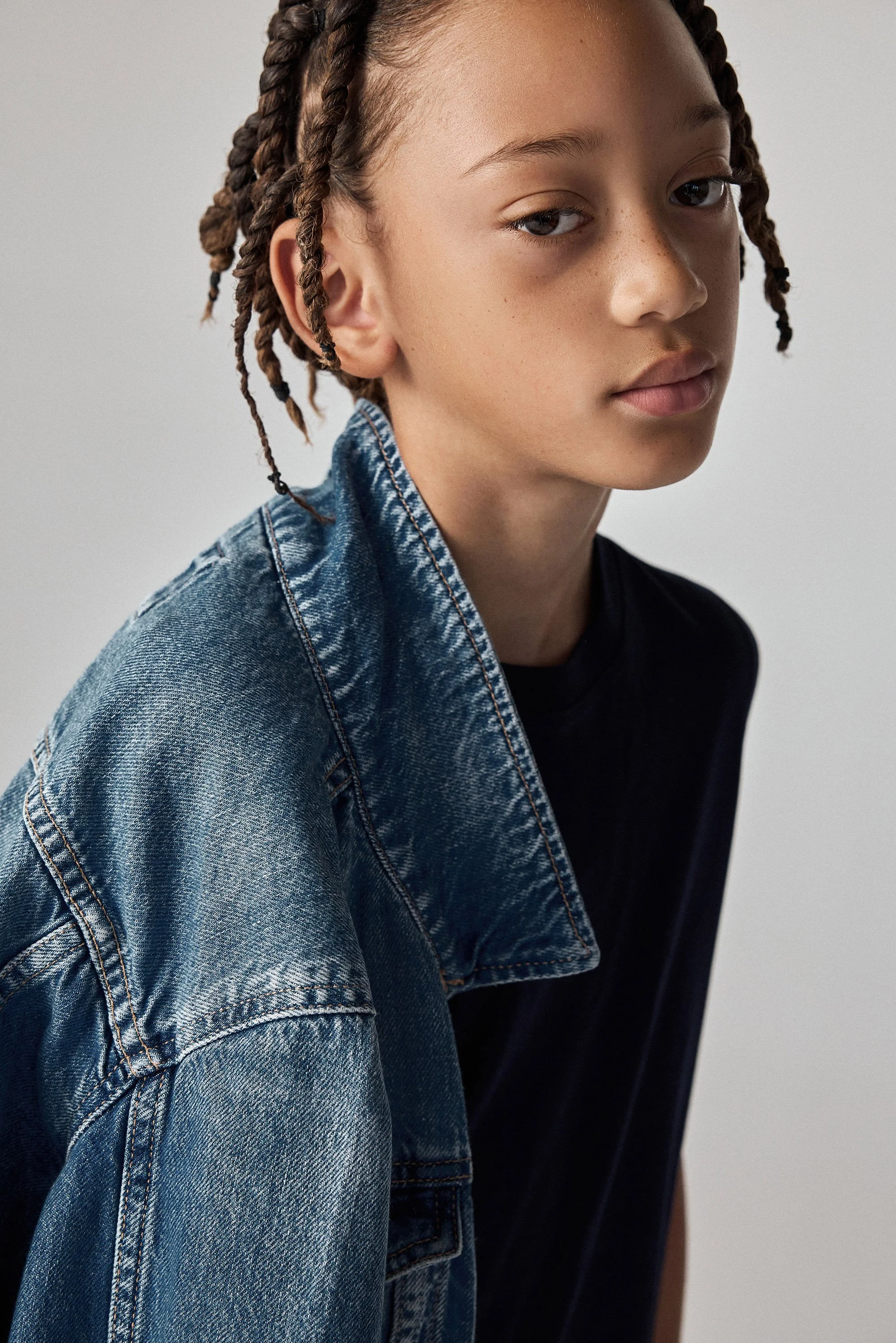 A young person with light brown skin and braided hair, wearing a denim jacket and a black shirt, looking at the camera with a neutral expression.