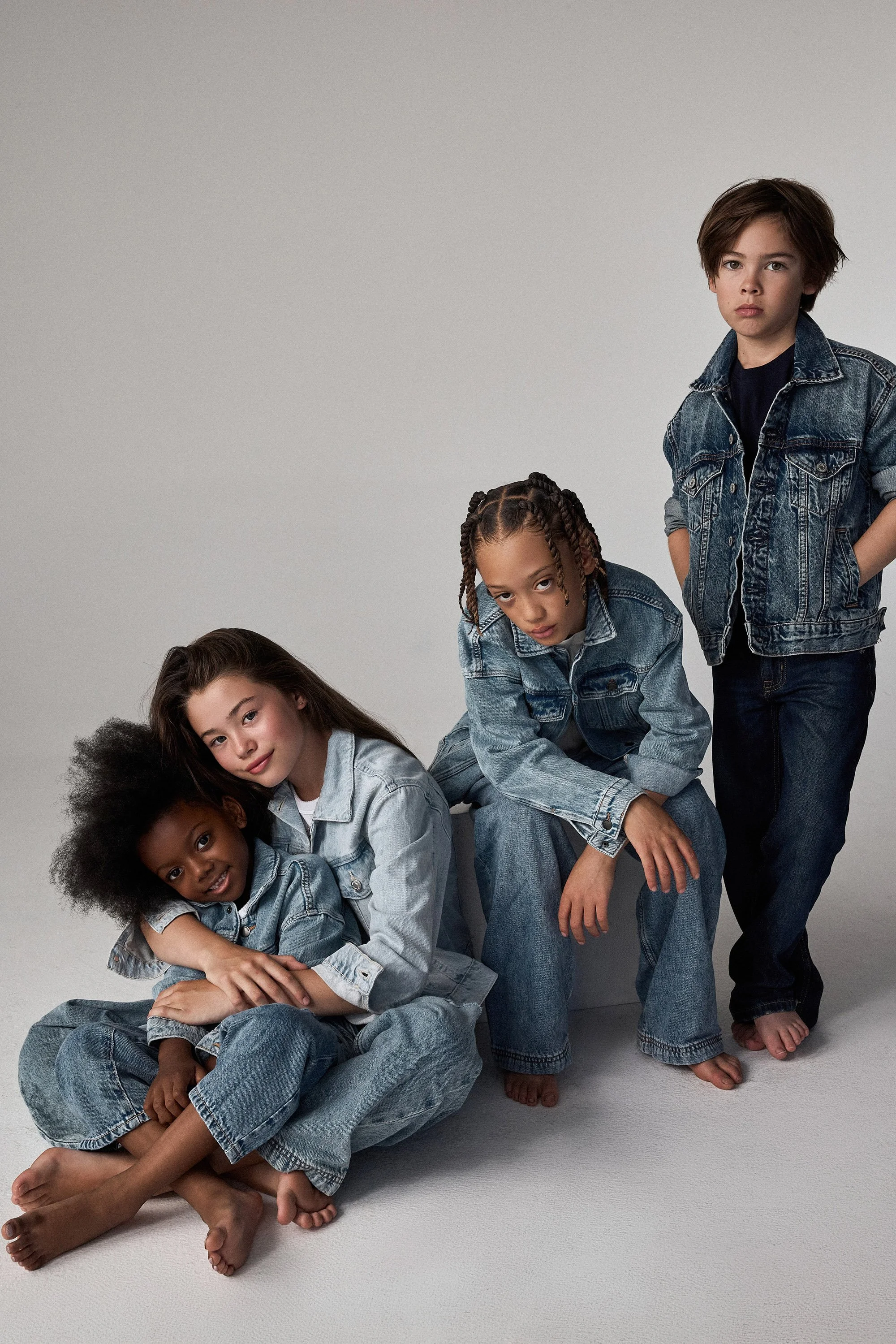 Four children wearing denim jackets and jeans posing against a plain background, with one seated on the floor, one leaning forward, and two standing.