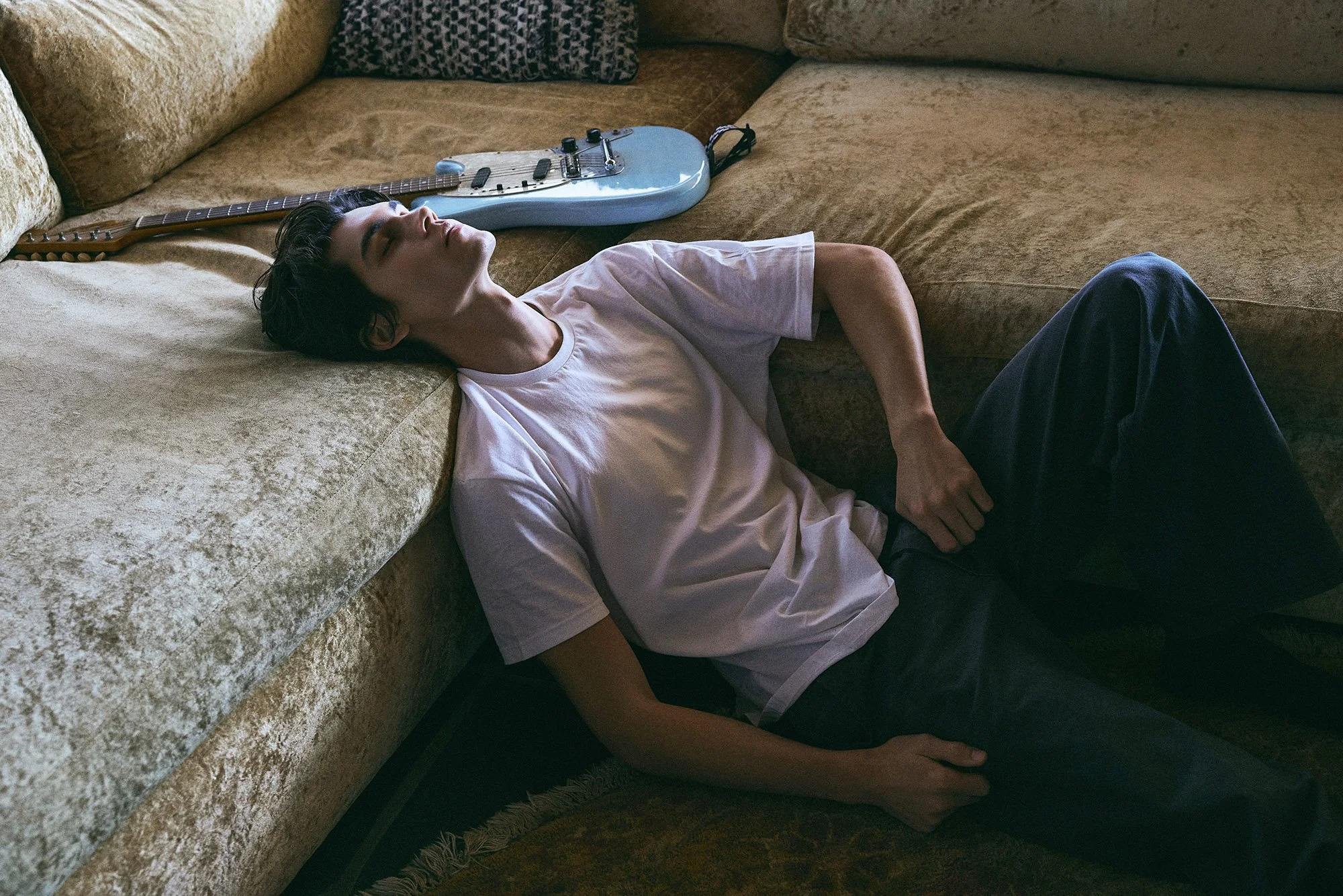 A young man with dark hair lying on a vintage yellow velvet couch with his head resting on the armrest. He is wearing a white t-shirt and dark pants. An electric guitar is lying next to him on the couch.