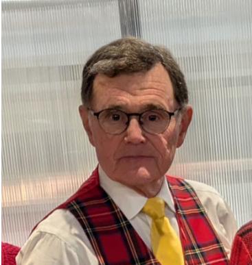 A portrait of an older man with glasses, wearing a blue suit, white shirt, and red tie, in front of a wood-paneled wall.