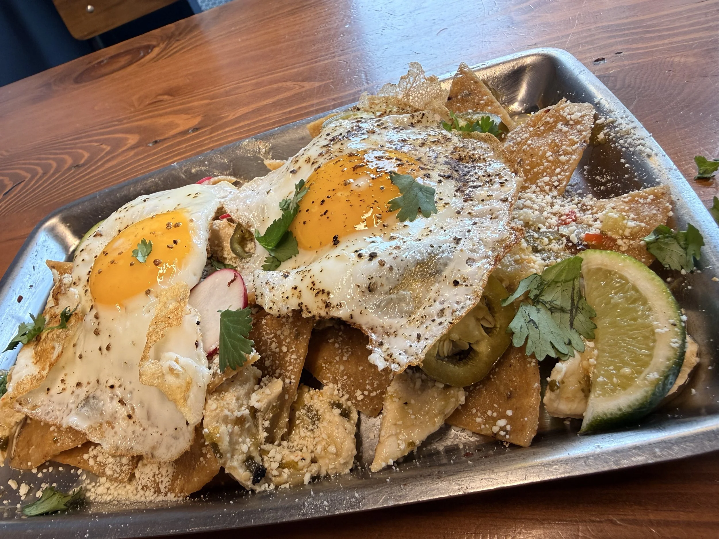 A tray of Mexican chilaquiles with fried eggs on top, garnished with cilantro, served with lime wedges, radish slices, and cilantro leaves