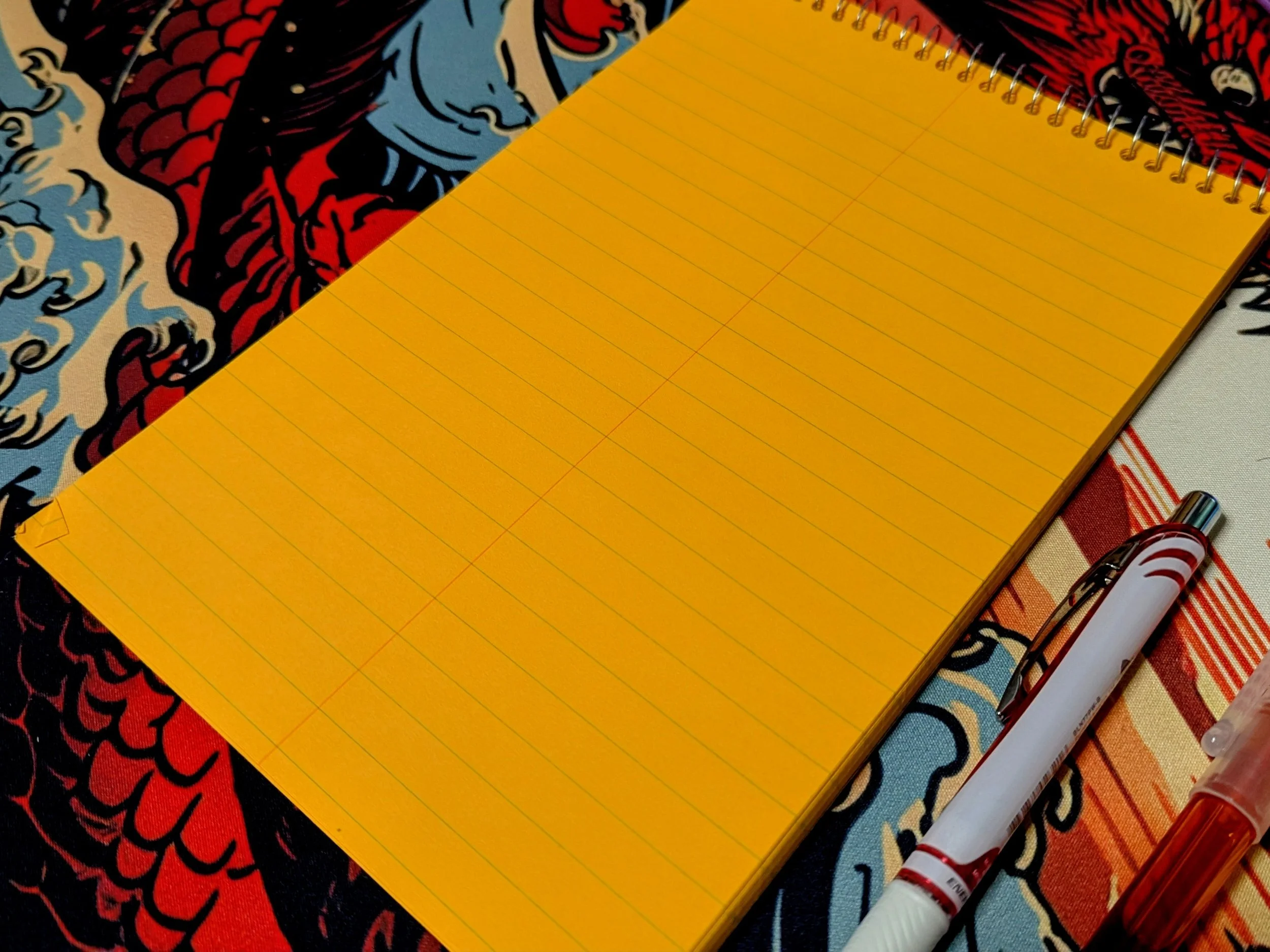 A notepad that is spiral bound with metal at the top that has blank, bright orange paper with printed ruled lines on it. It sits diagonally on a very colorful background with a high contrast dragon design and has two pens lying to the right of it.