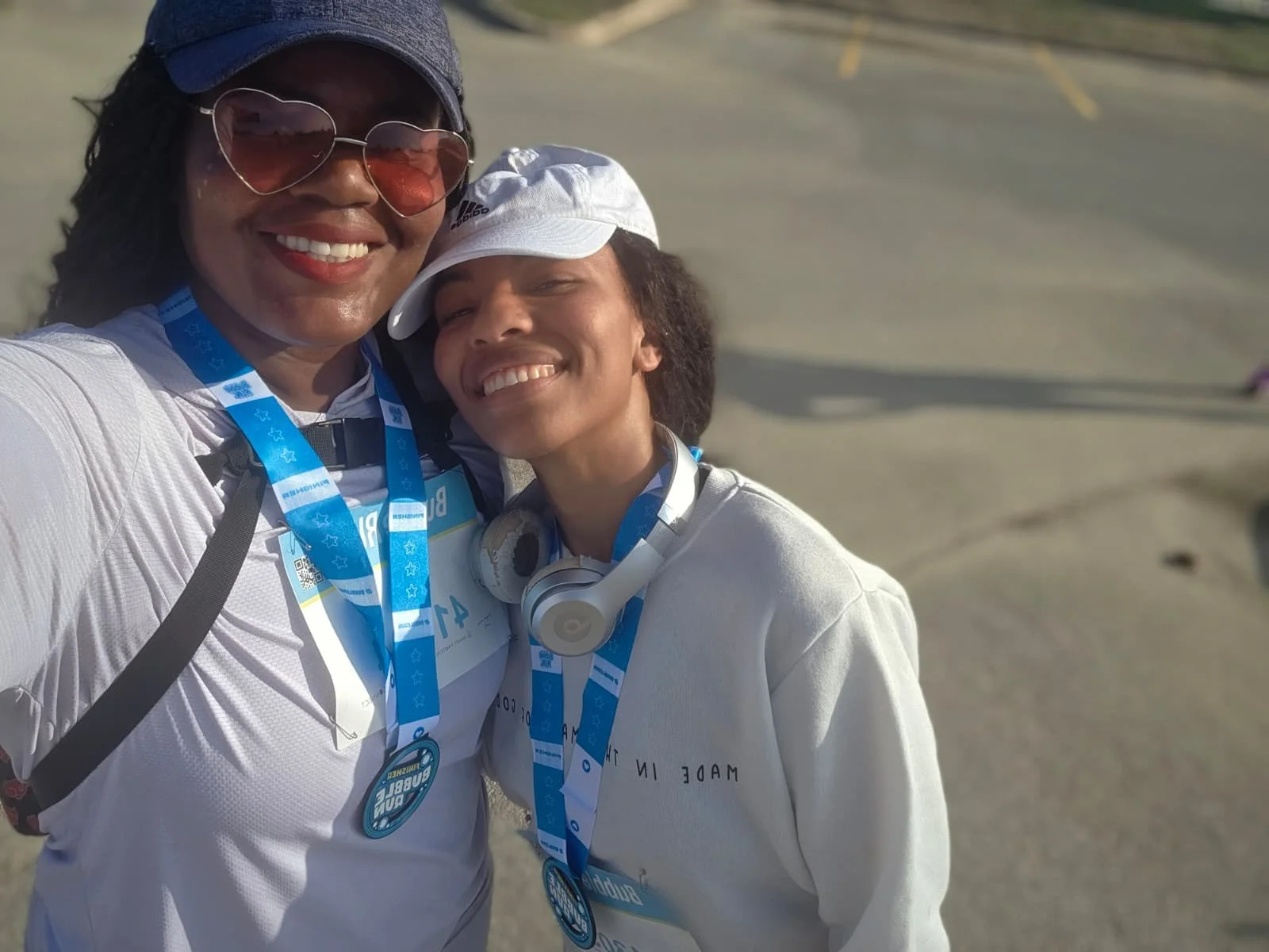 Two women with medals around their necks taking a selfie outdoors, smiling at the camera. One woman is wearing sunglasses and a gray cap, while the other has headphones around her neck and a white cap.