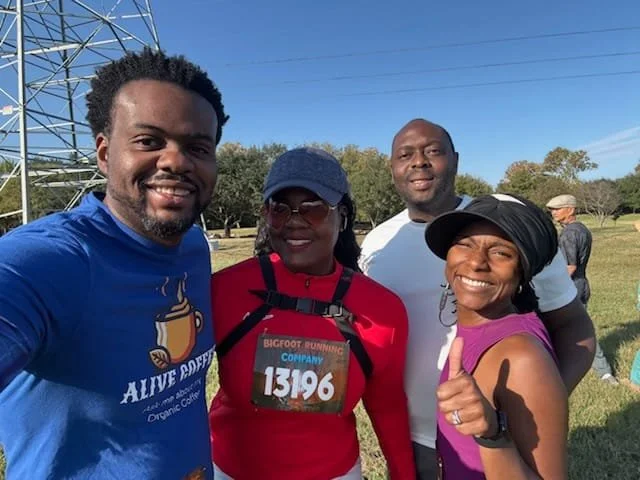 Four people standing outdoors, smiling, with a field and power lines in the background. One person is taking a selfie. The group includes two women and two men, dressed casually, with one woman wearing a race bib that says 13196.