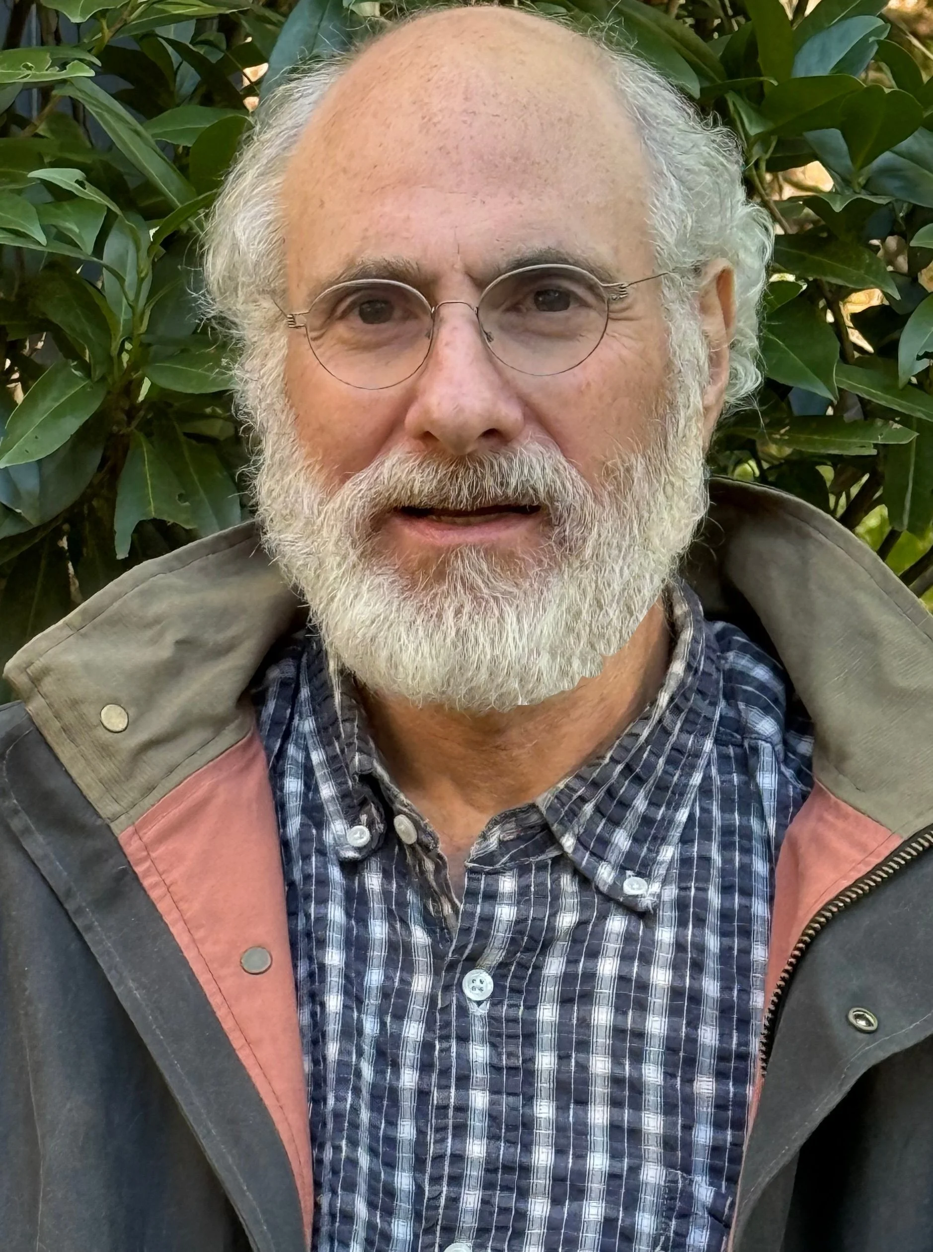 Close-up of an older man with glasses, white beard, and curly hair, standing outdoors in front of green foliage.