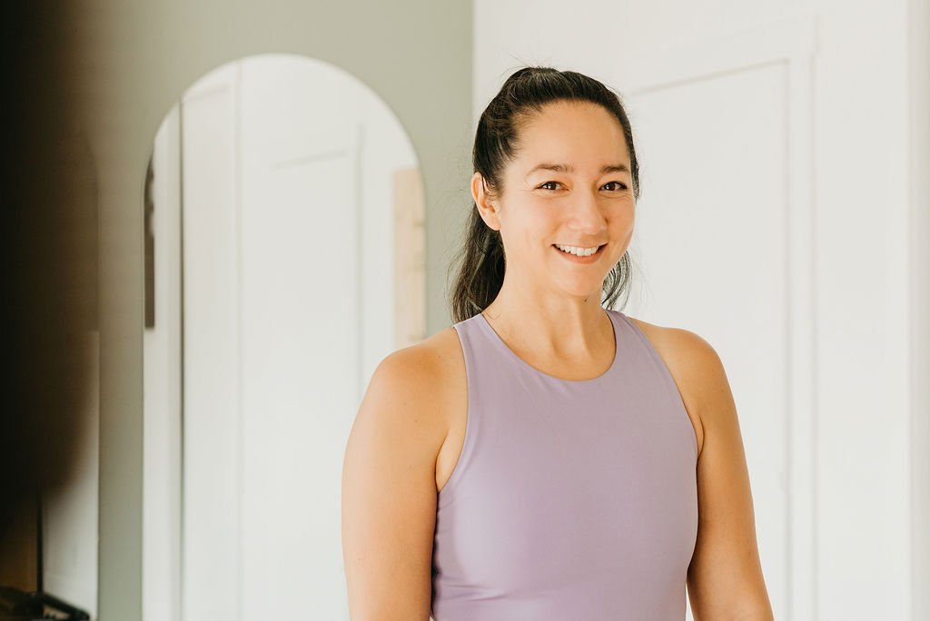 Smiling woman with dark hair tied back, wearing a light purple sleeveless athletic top, standing indoors near a mirror.