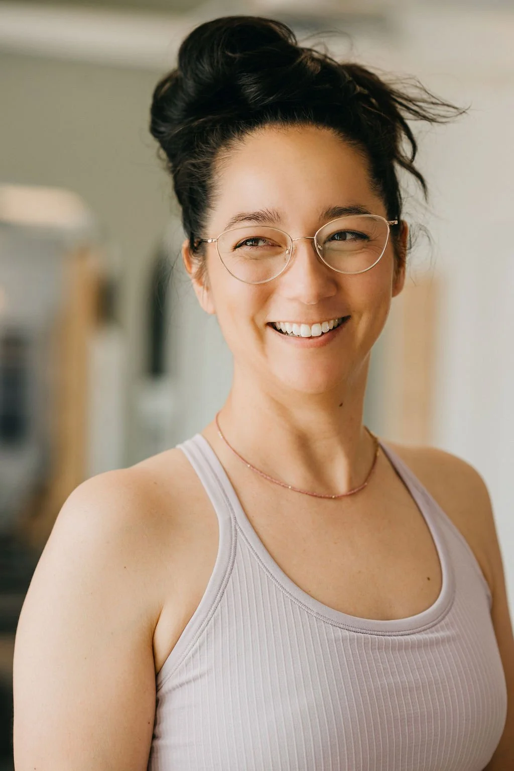 A woman with dark hair styled in a messy bun, wearing glasses and a light-colored tank top, smiling warmly in an indoor setting.