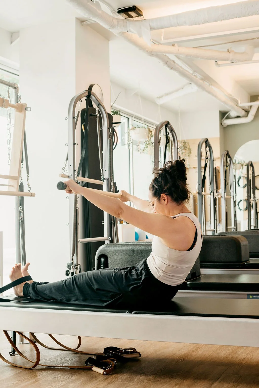 A woman doing pilates on a reformer machine in a brightly lit gym with multiple reformer machines.