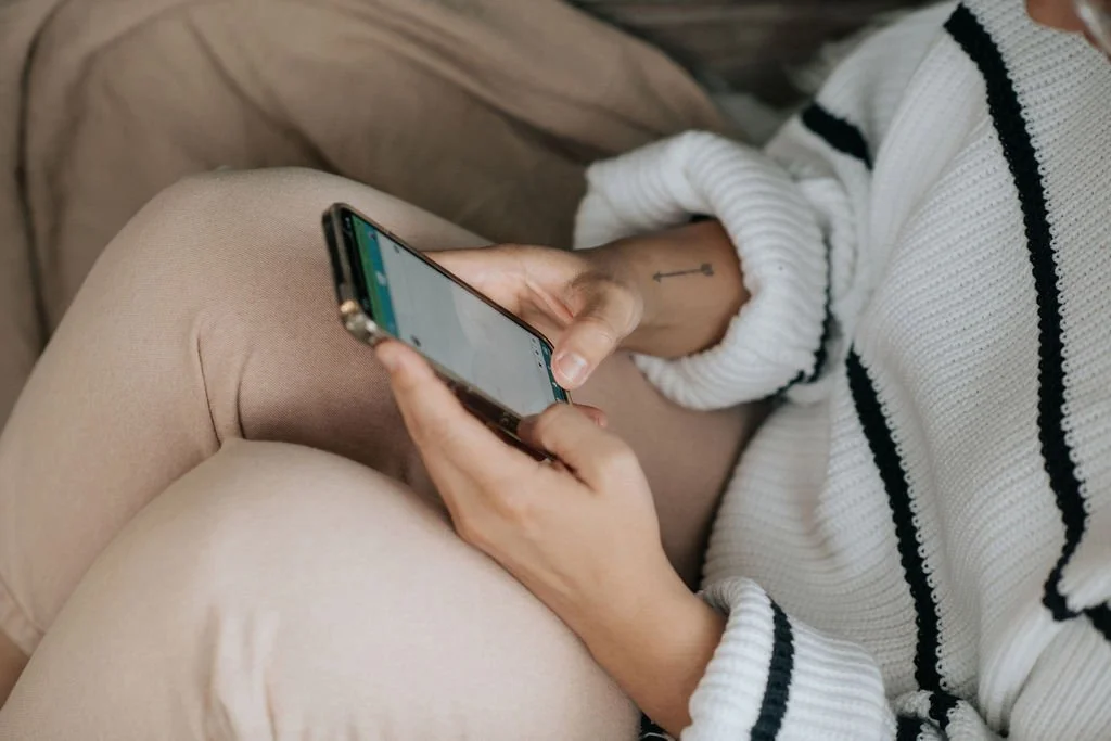 Person sitting cross-legged, wearing beige pants and a white sweater with black stripes, holding and using a smartphone.