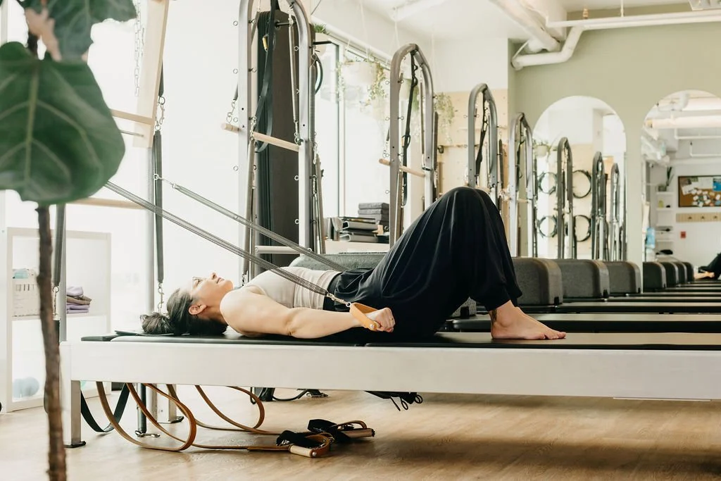 A woman lying on her back on a Pilates reformer, holding the handles with her arms, in a fitness studio with large windows and Pilates equipment in the background.