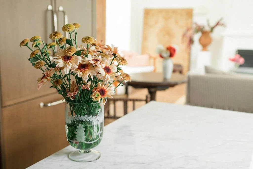 A glass vase with a bouquet of various flowers on a white table in a bright room.