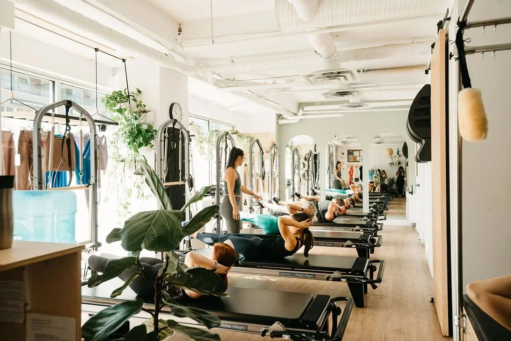 People participating in a Pilates class using reformer machines in a bright, airy studio with large windows and plants.