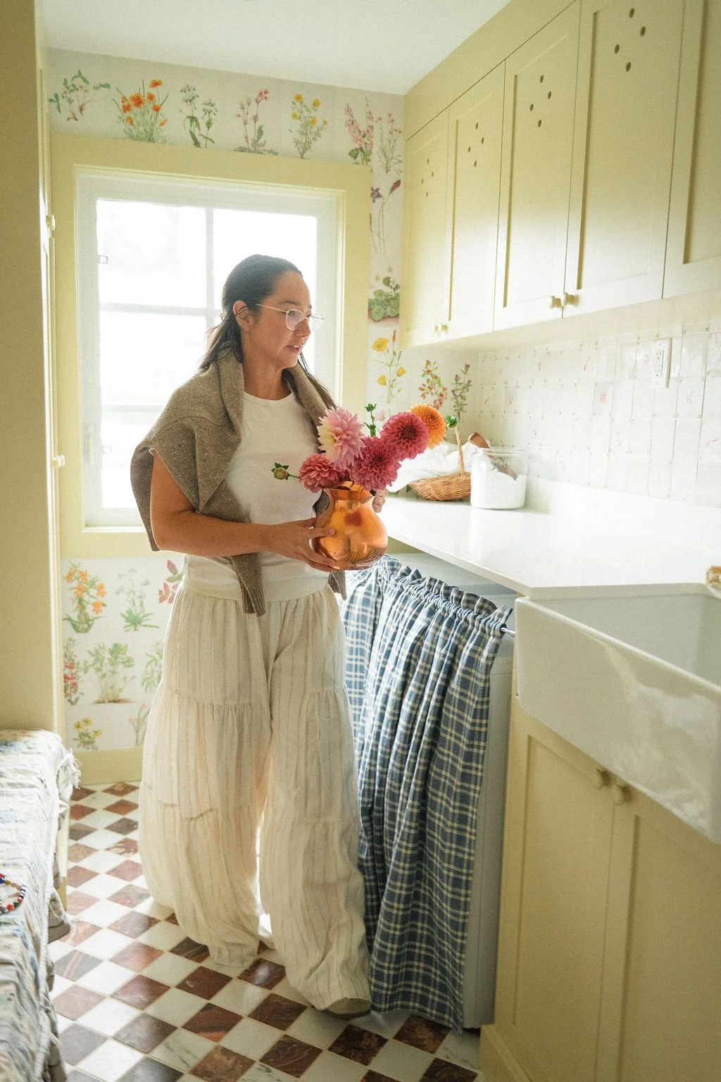 Woman holding a vase of pink and orange flowers in a bright, floral-themed kitchen with yellow cabinets and checkered linoleum floor.